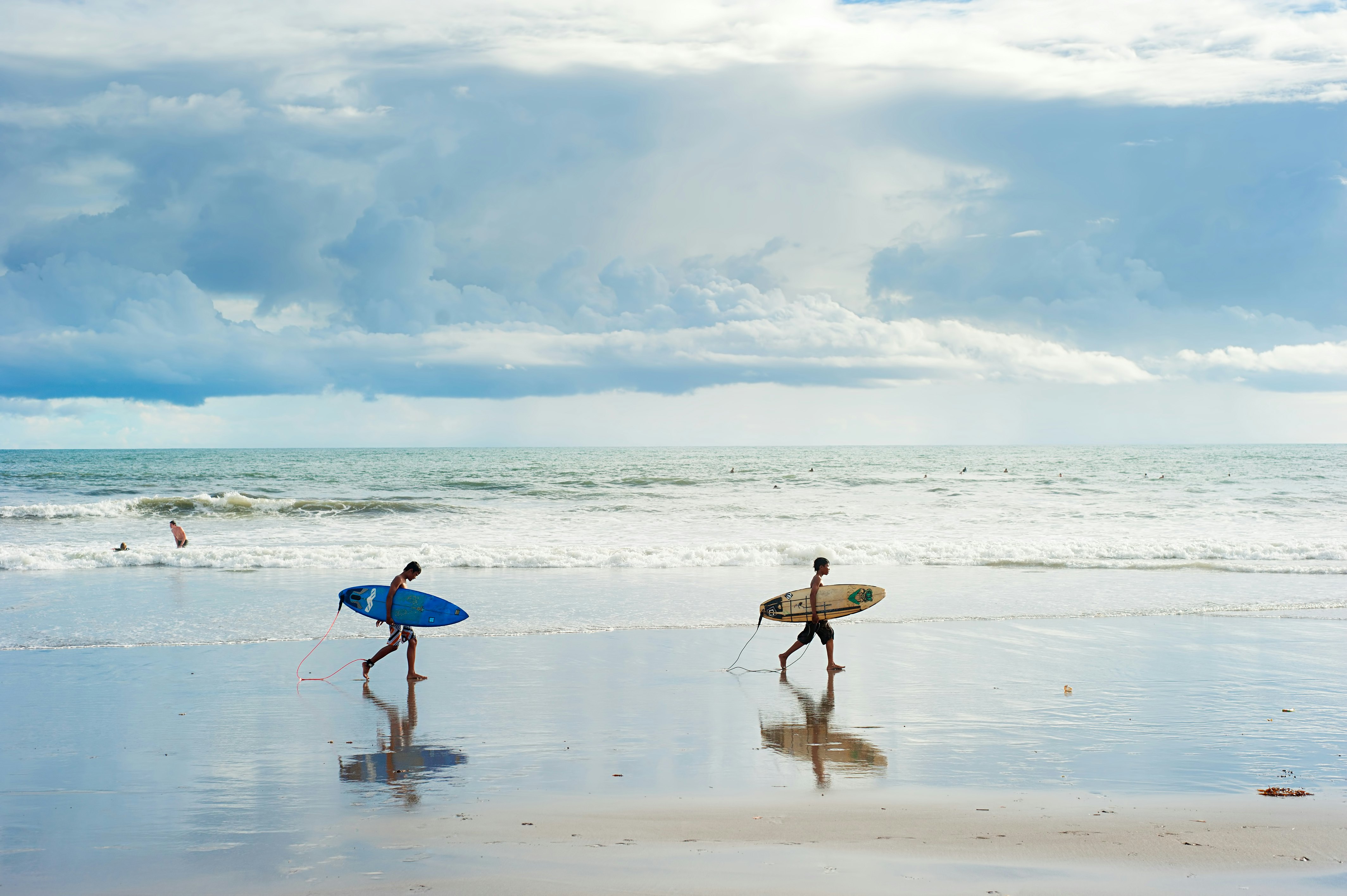 Two surfers walk with surfboards parallel to the water on a beach.
