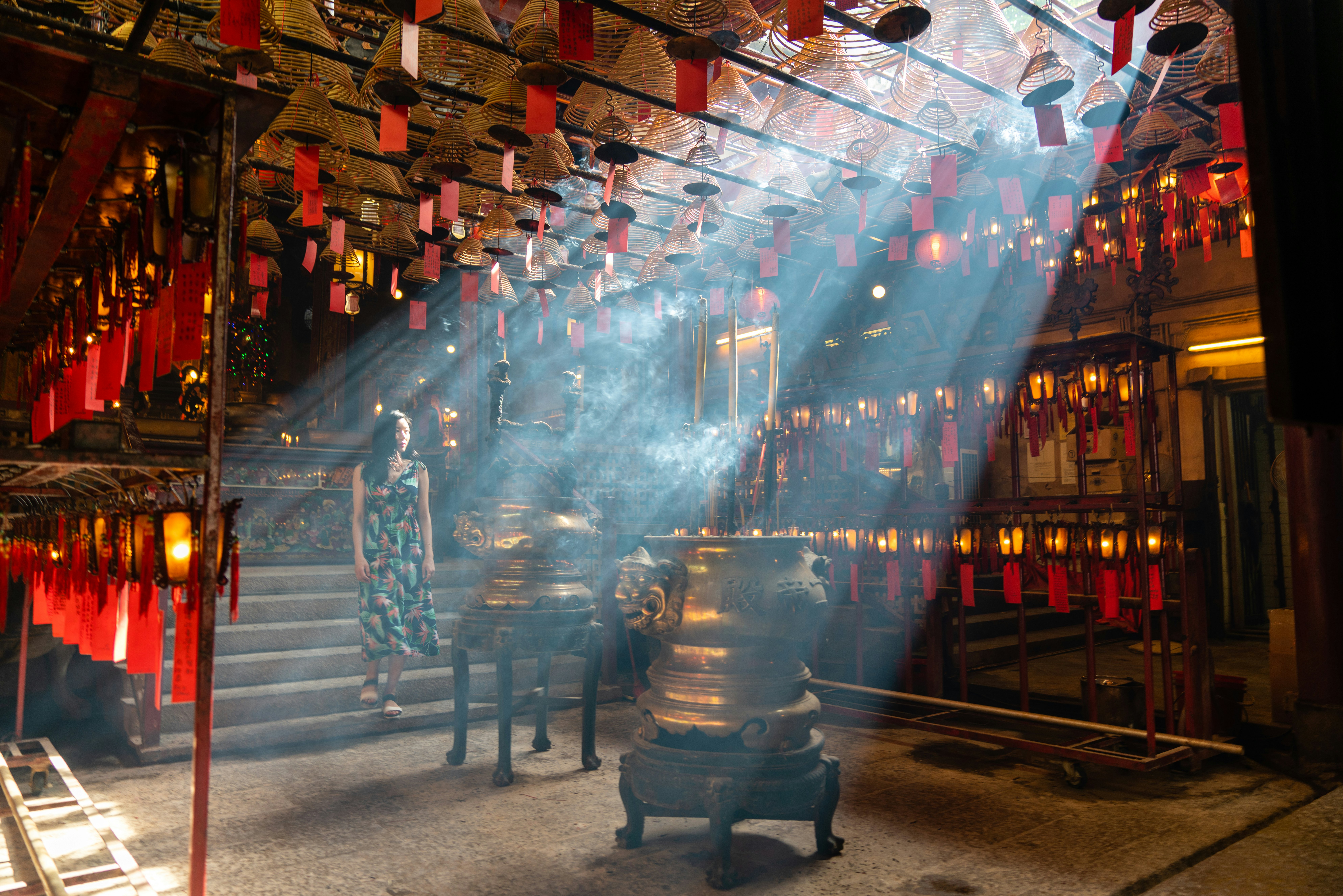 A woman in a floral dress walks down stairs in a temple, with lanterns to the side and large vessels in the middle. Light streams through an upper window.