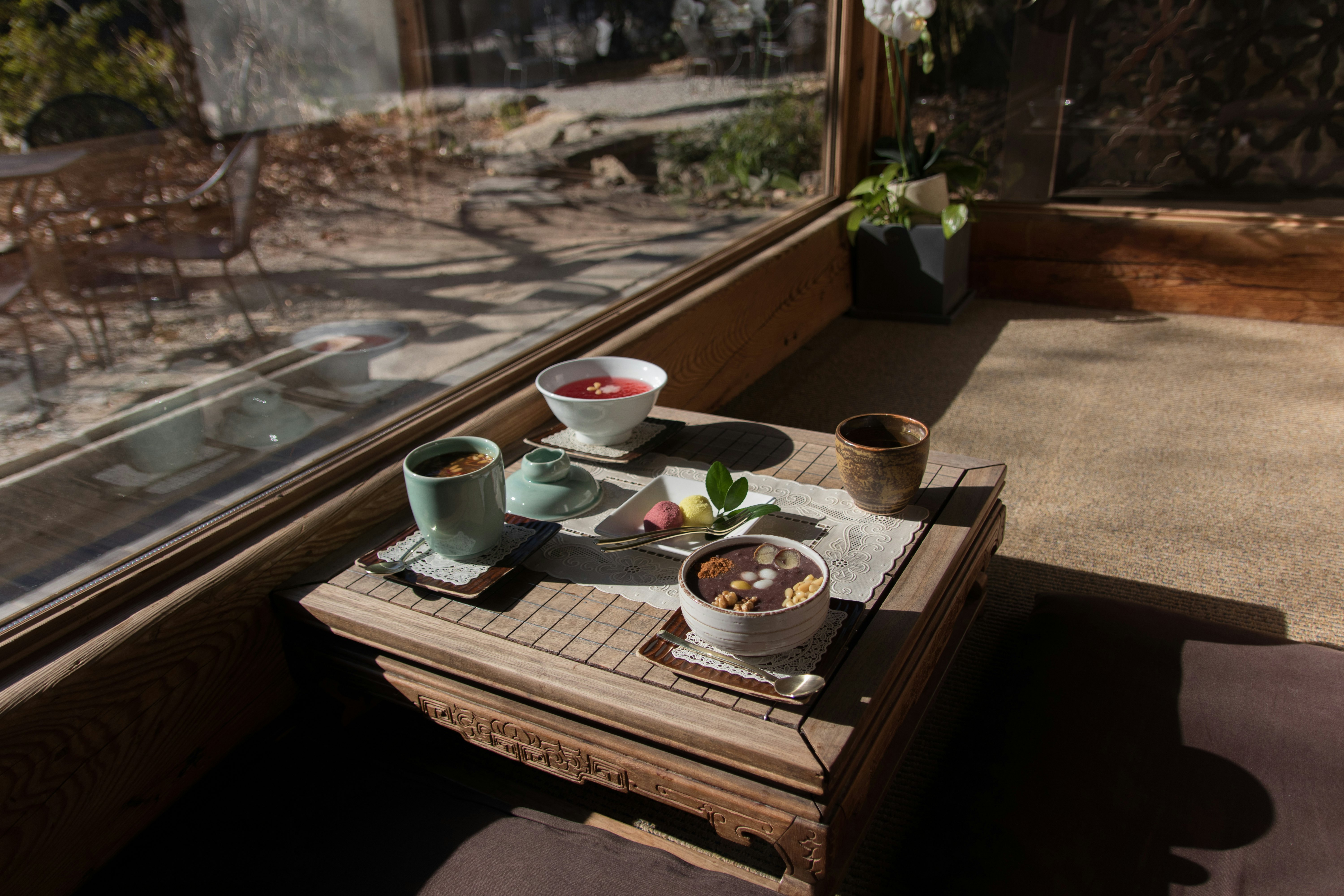 Cups and bowls are carefully set on a small table in a teahouse. A small garden is seen through a large window next to the table.