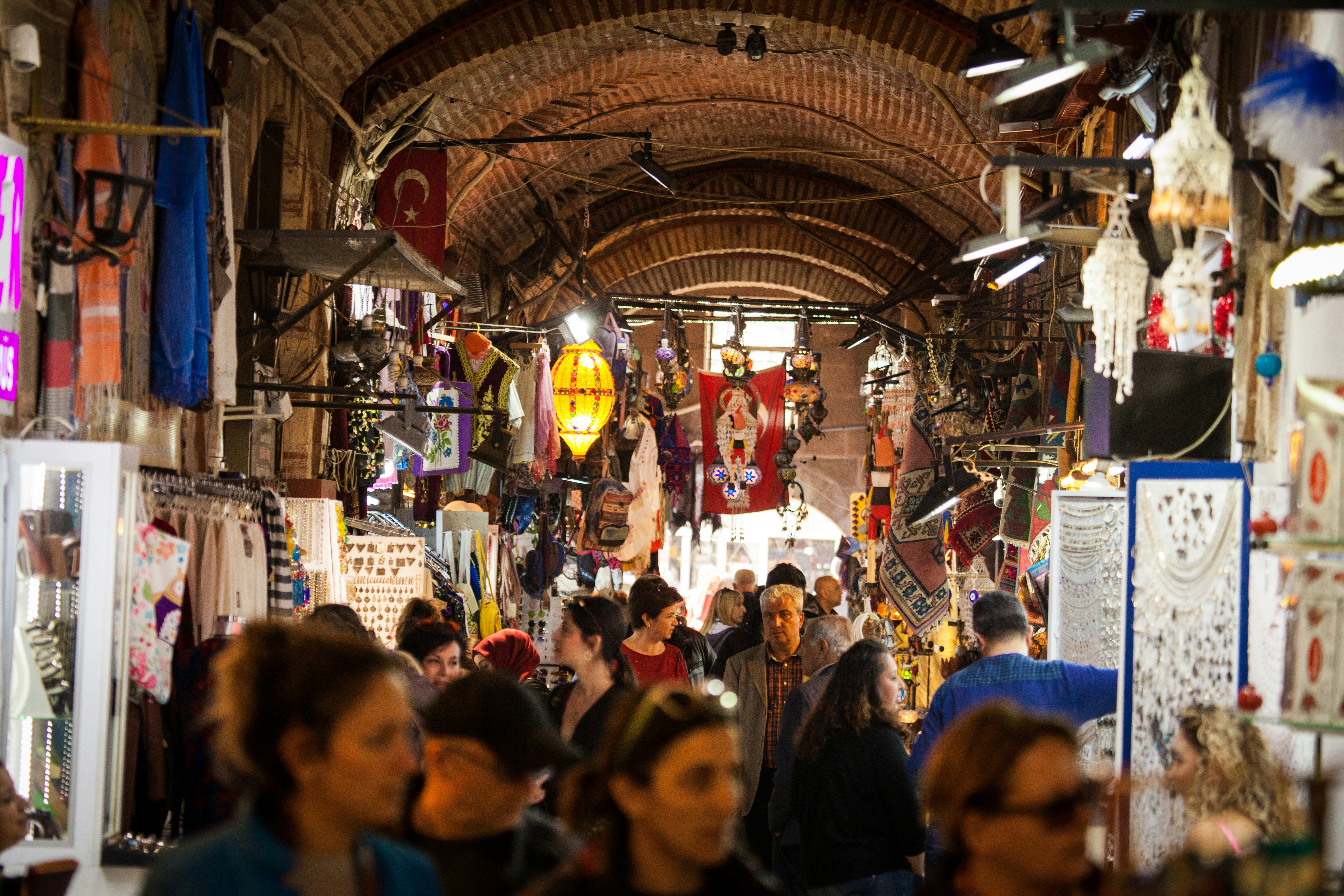 A crowd walks through an arched passageway of a bazaar, with vendors selling jewelry, lighting fixtures and other items.