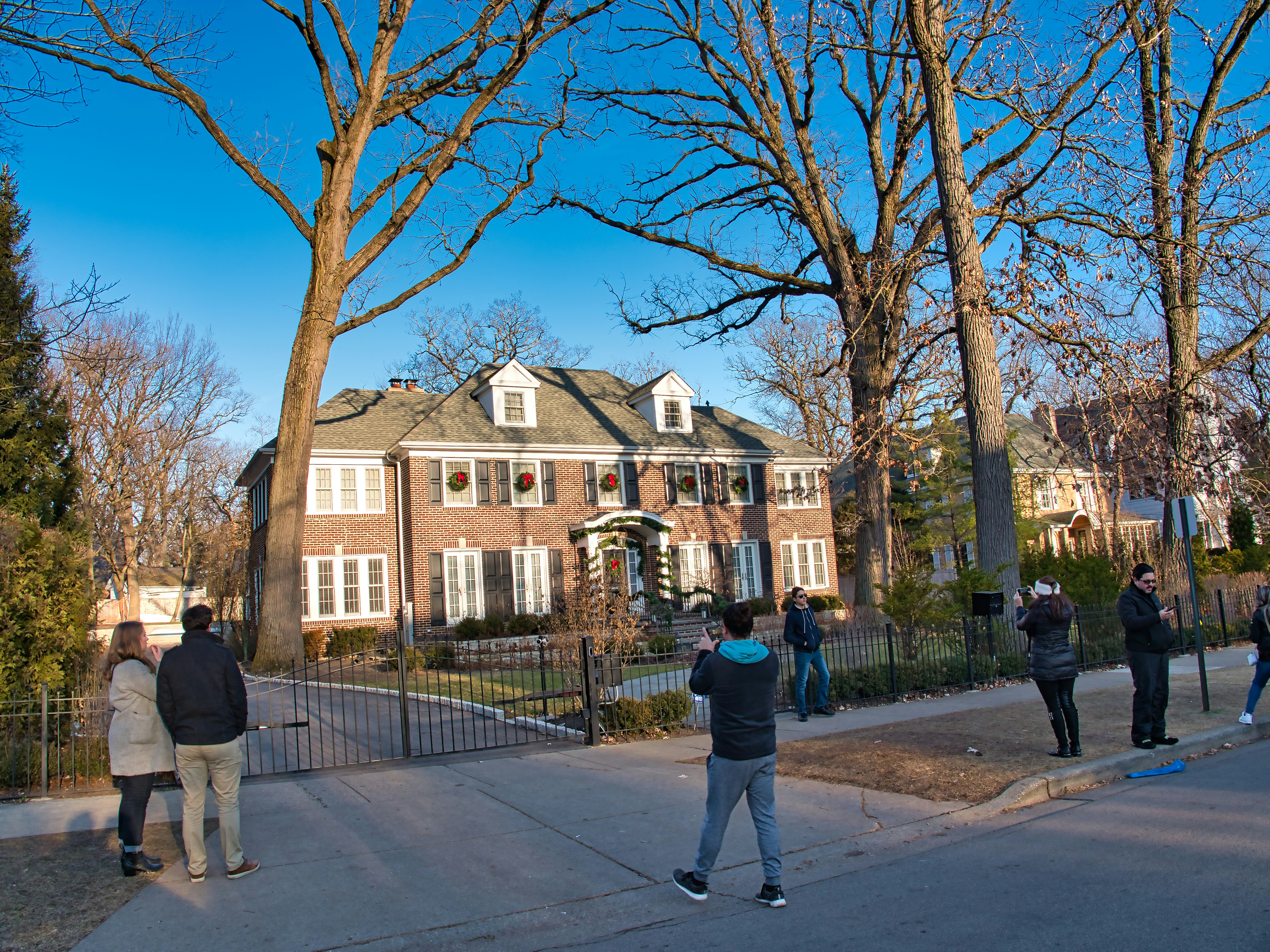 People gather to take pictures of a brick house behind a metal fence on a suburban street.