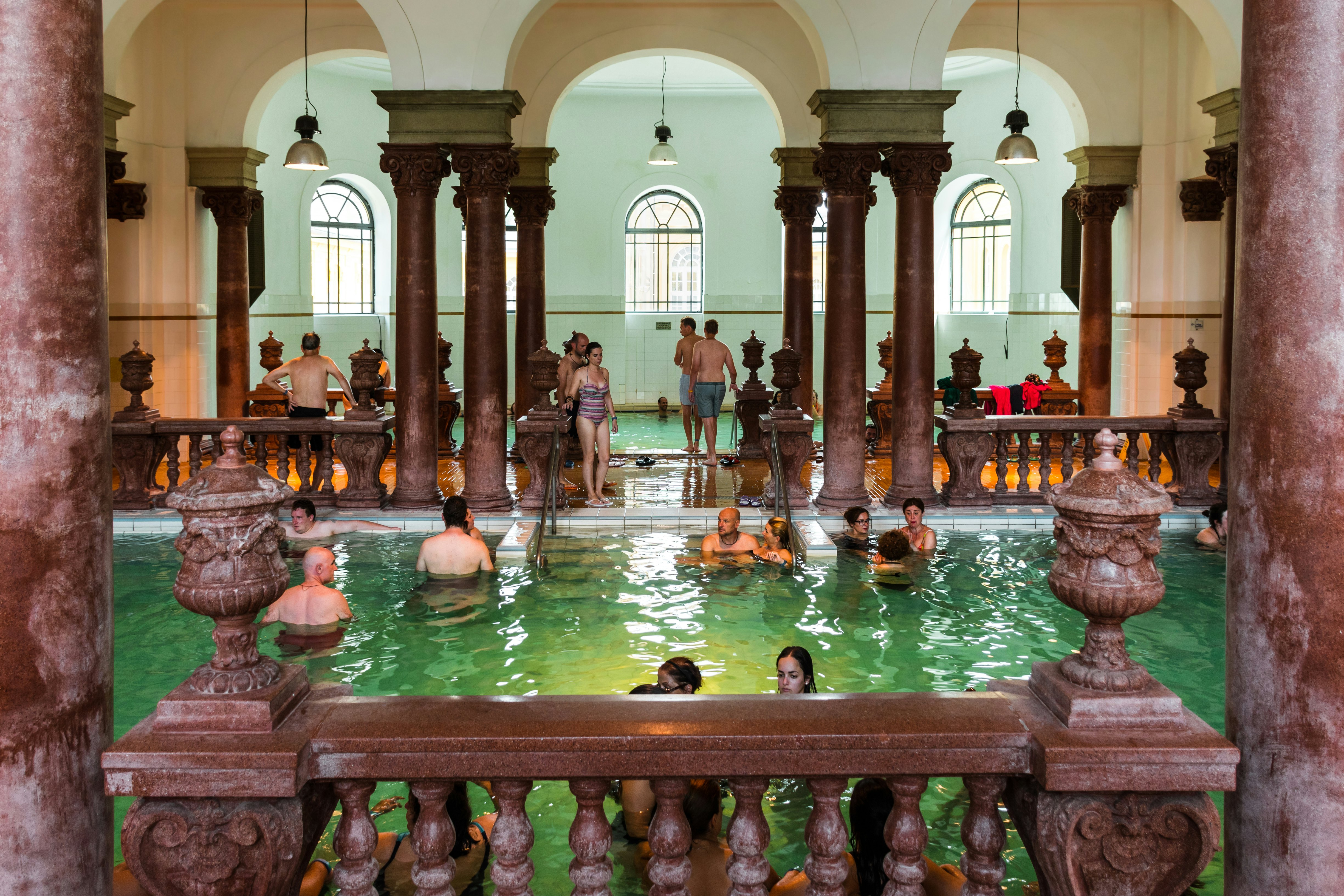 People are pictured in an around an indoor pool at thermal bath. Ornate columns and balustrades surround the pool.