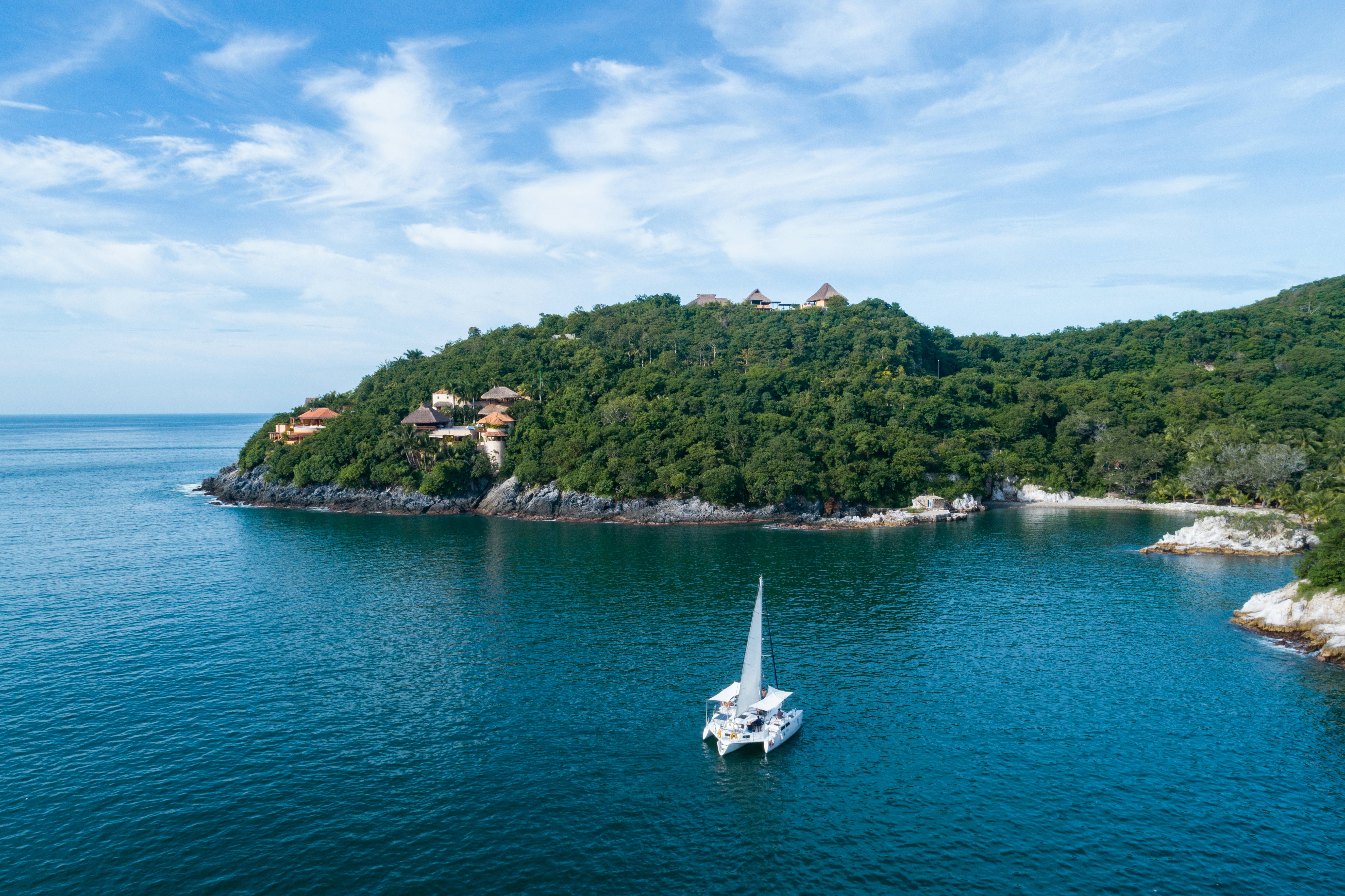 A wide view of a catamaran in a bay, near a shore with lushly forested hills.