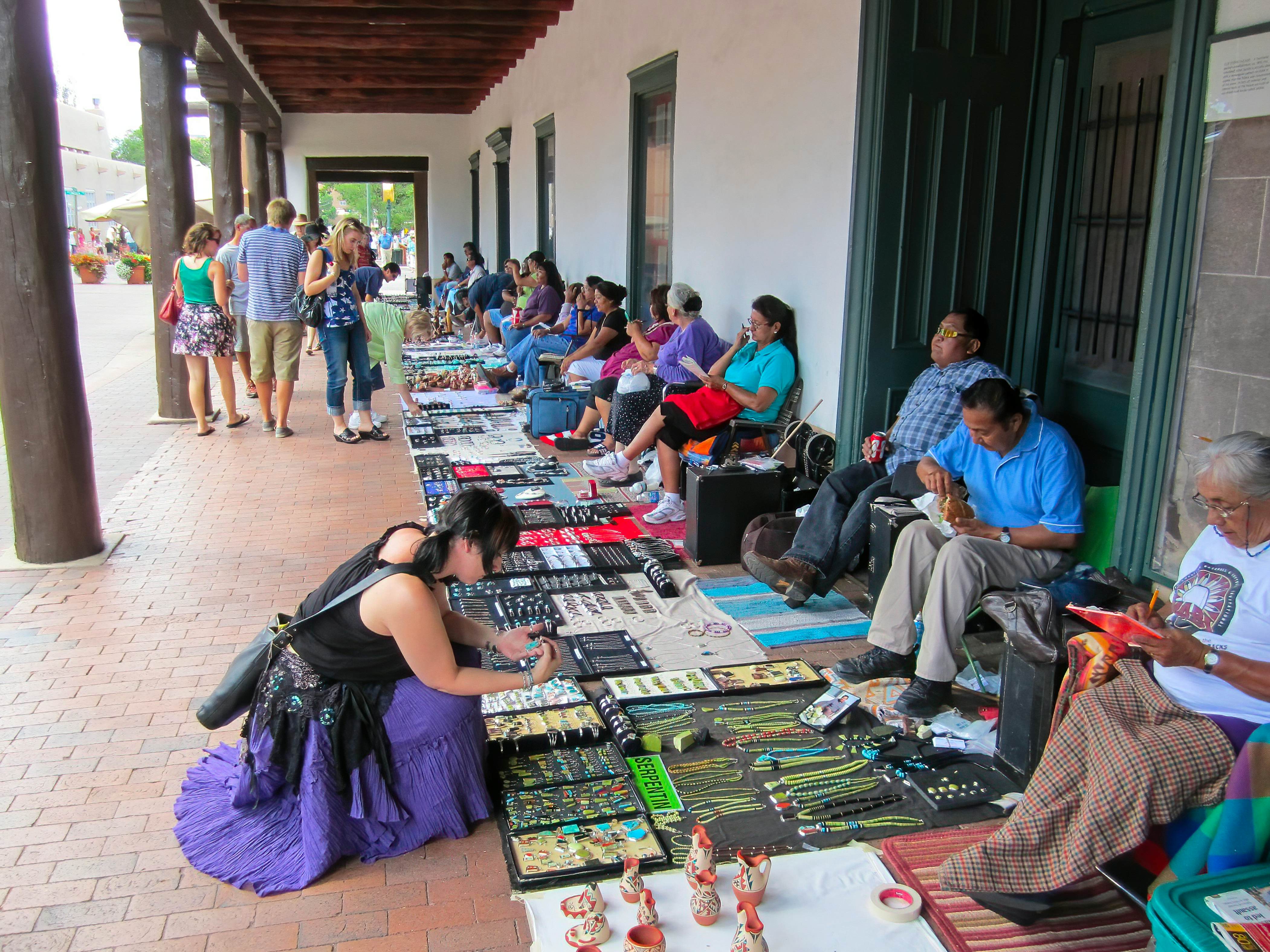 Shoppers look at goods spread on blankets on a walkway under an arcade. Vendors sit in chairs on the other side.