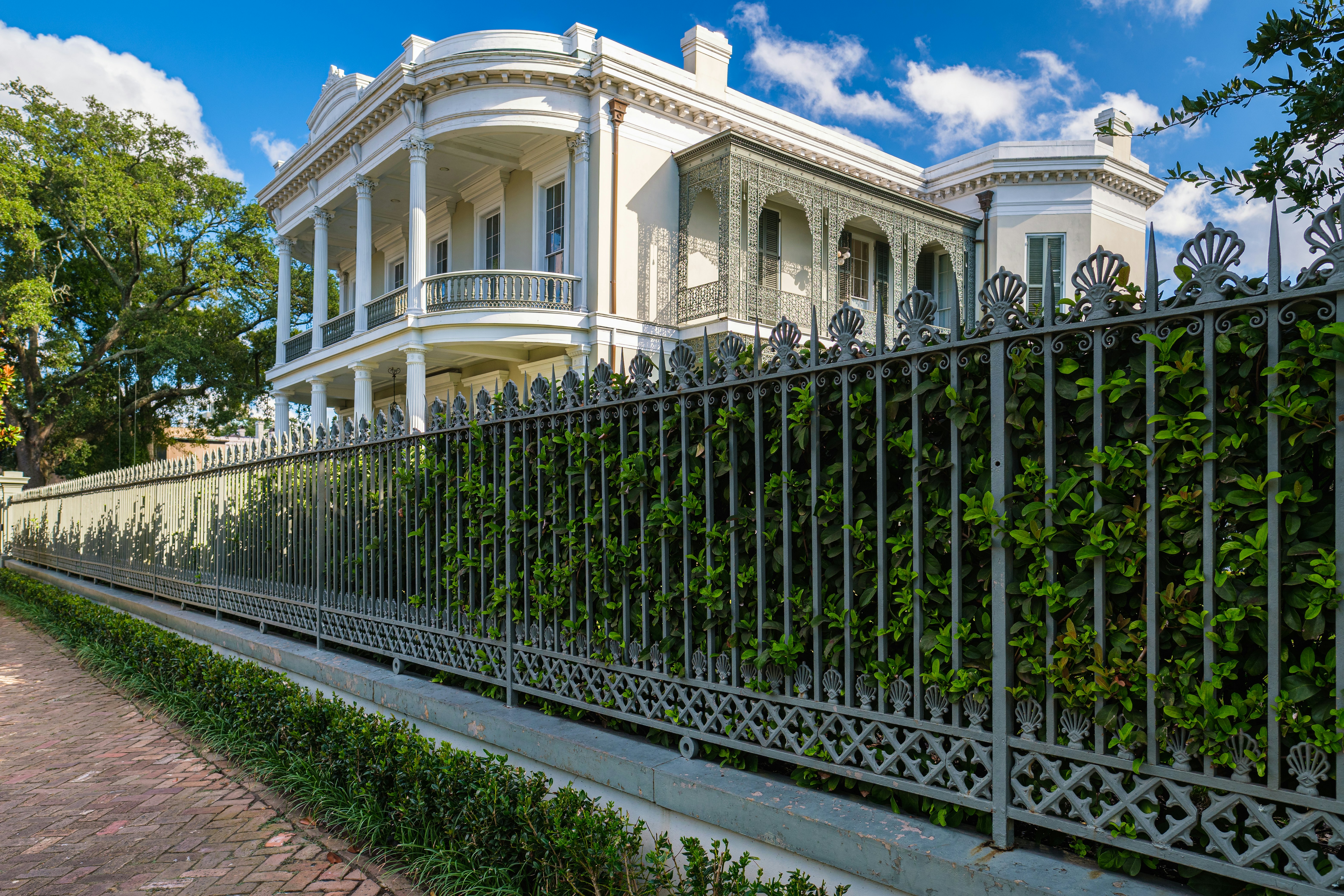 A shrub-lined metal fence in front of a large old beige mansion