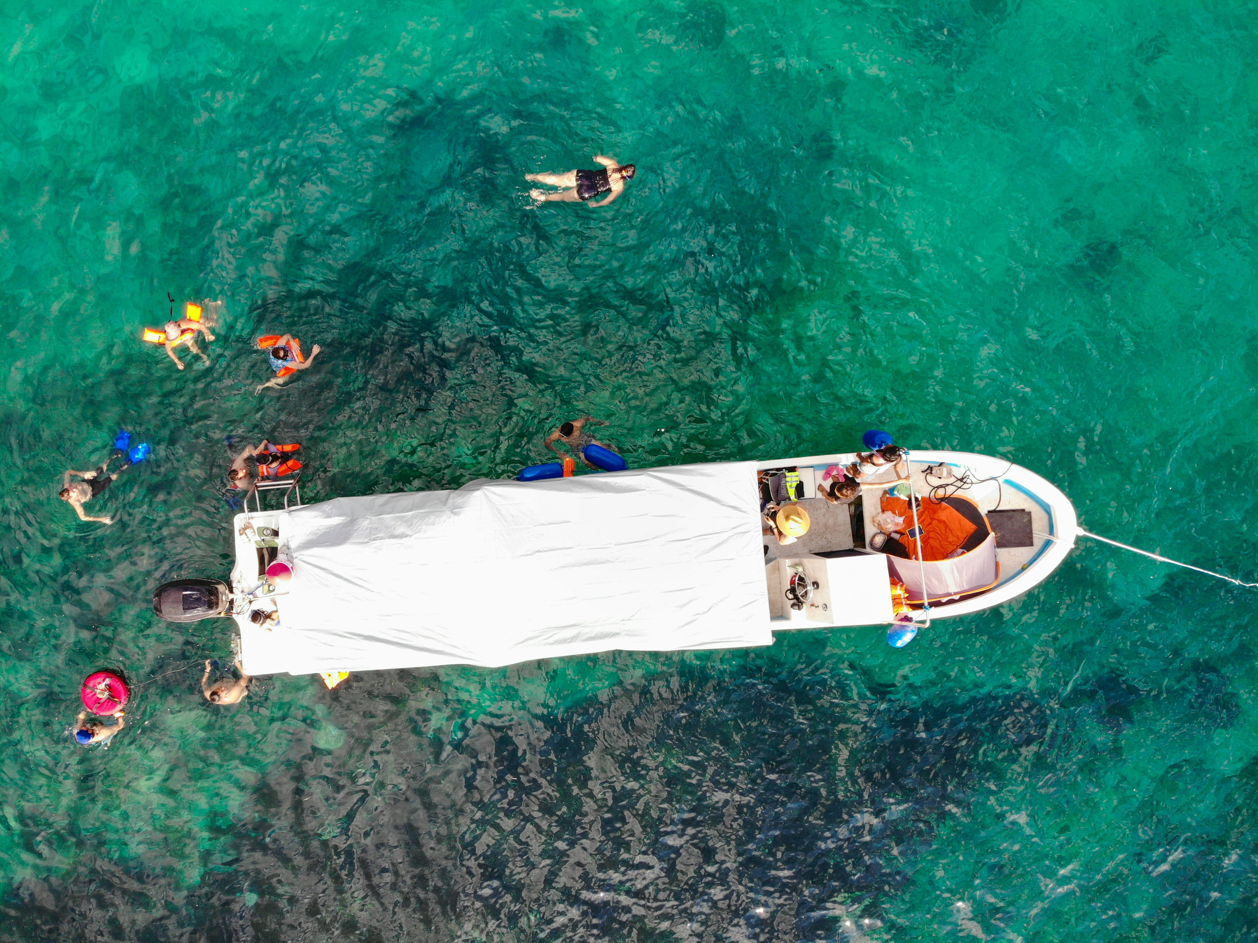 An overhead view of a small boat with a sun cover. People snorkel and swim in the clear water next to the boat.