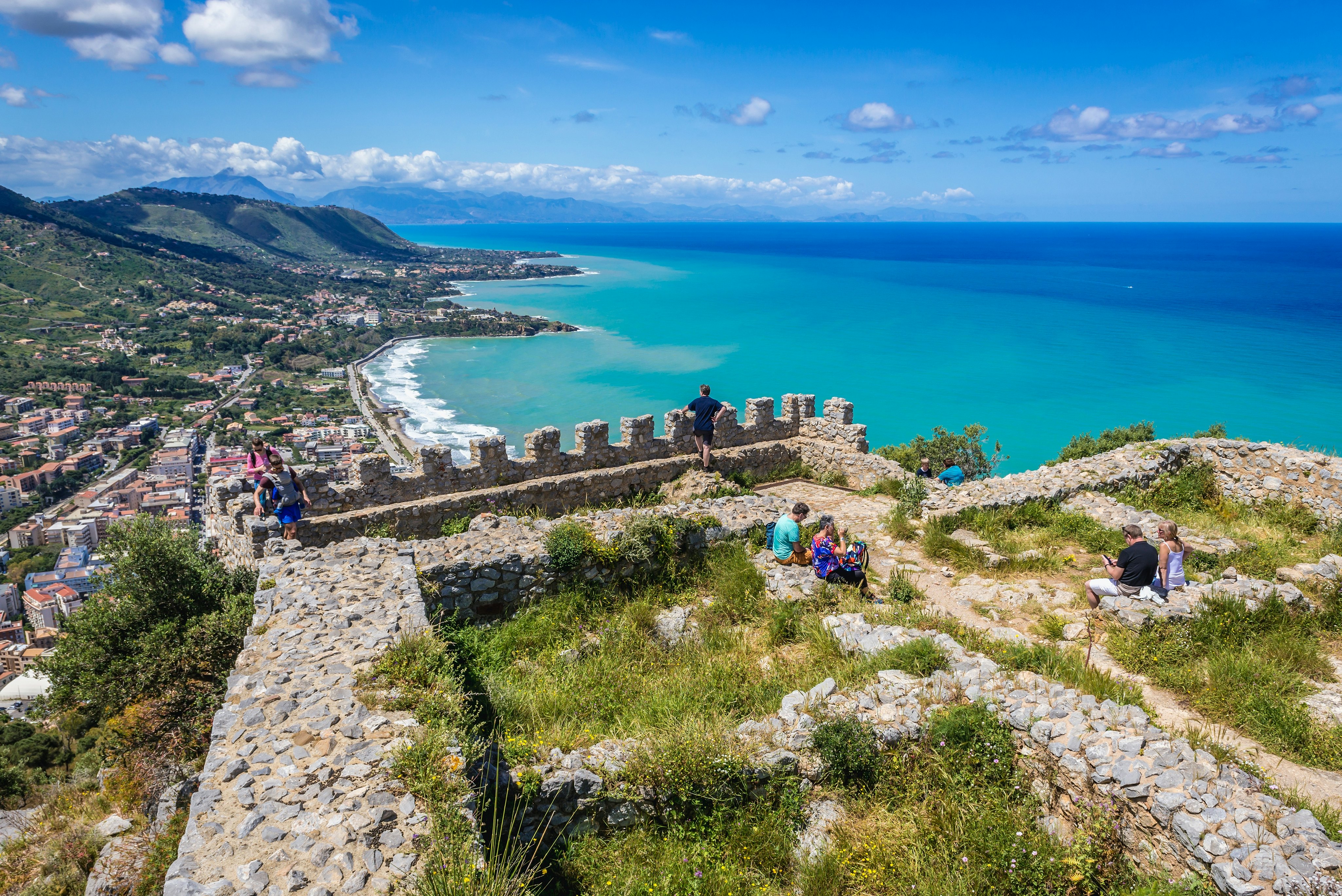 Castle ruins at the top of a hill overlooking a coastal city. Hikers rest on the stone walls.