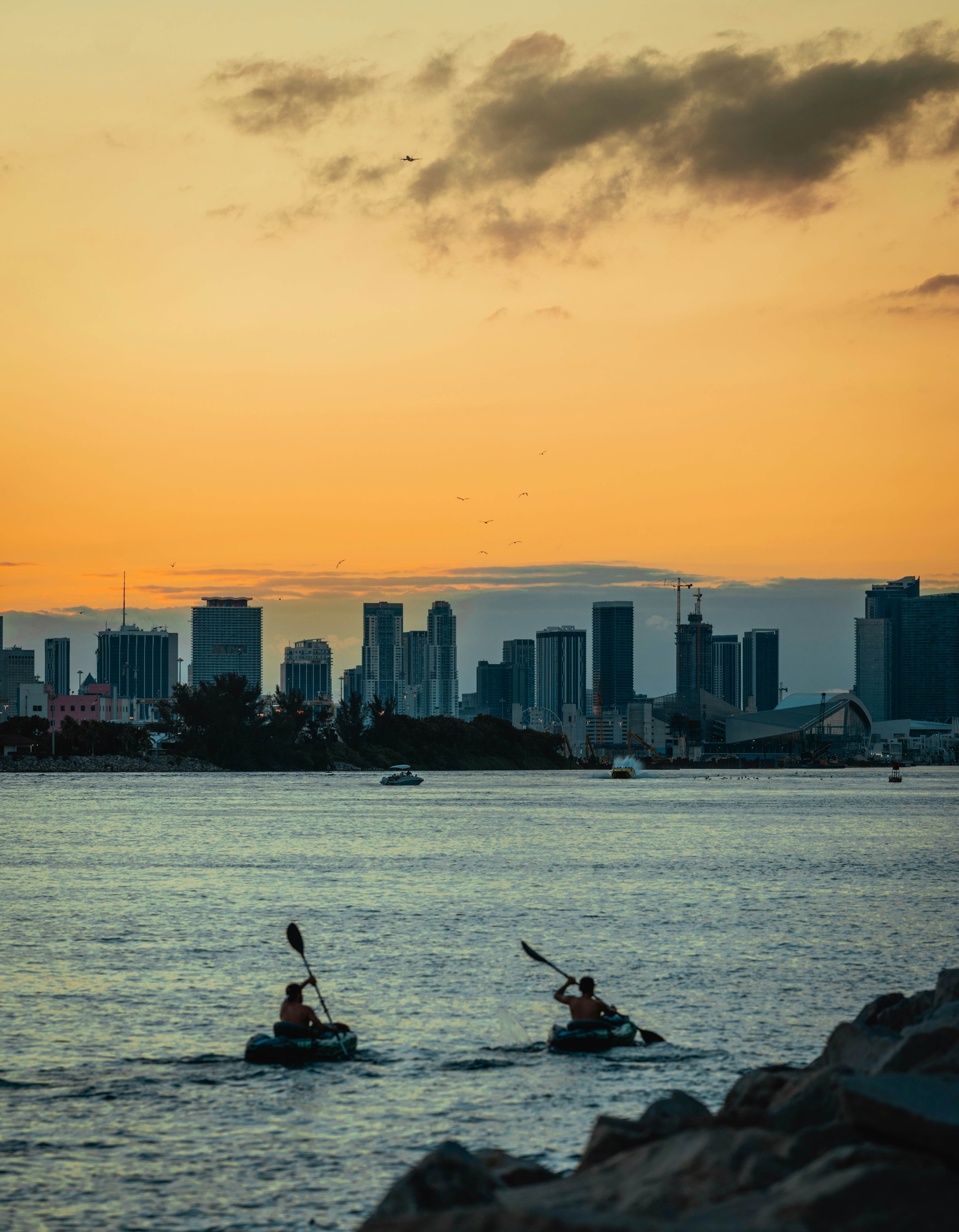 Two kayakers in silhouette as they paddle in a waterway near skyscrapers at sunset. 