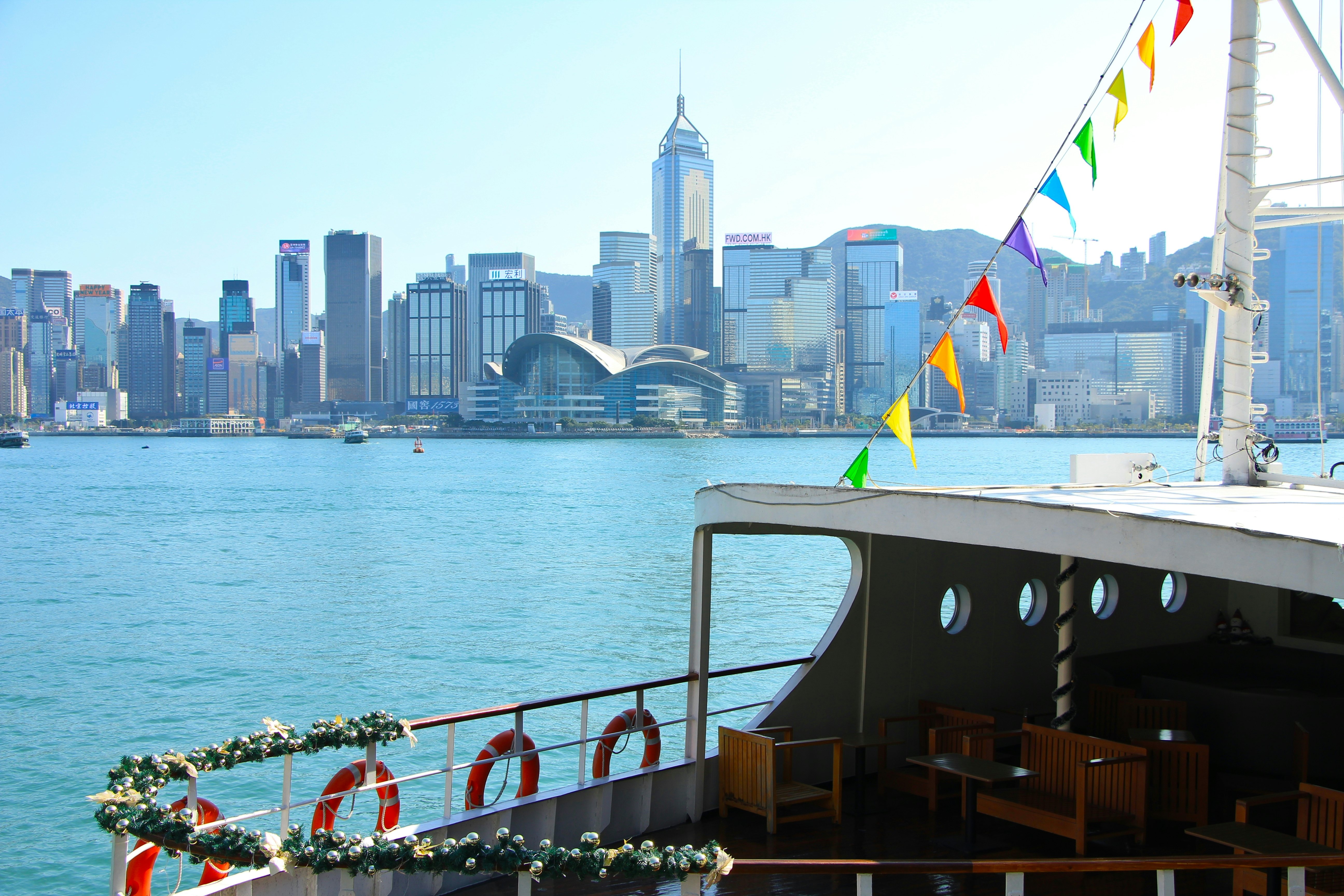 The upper deck of a ferry has multicolored bunting attached to the mast; a city skyline is across the water.