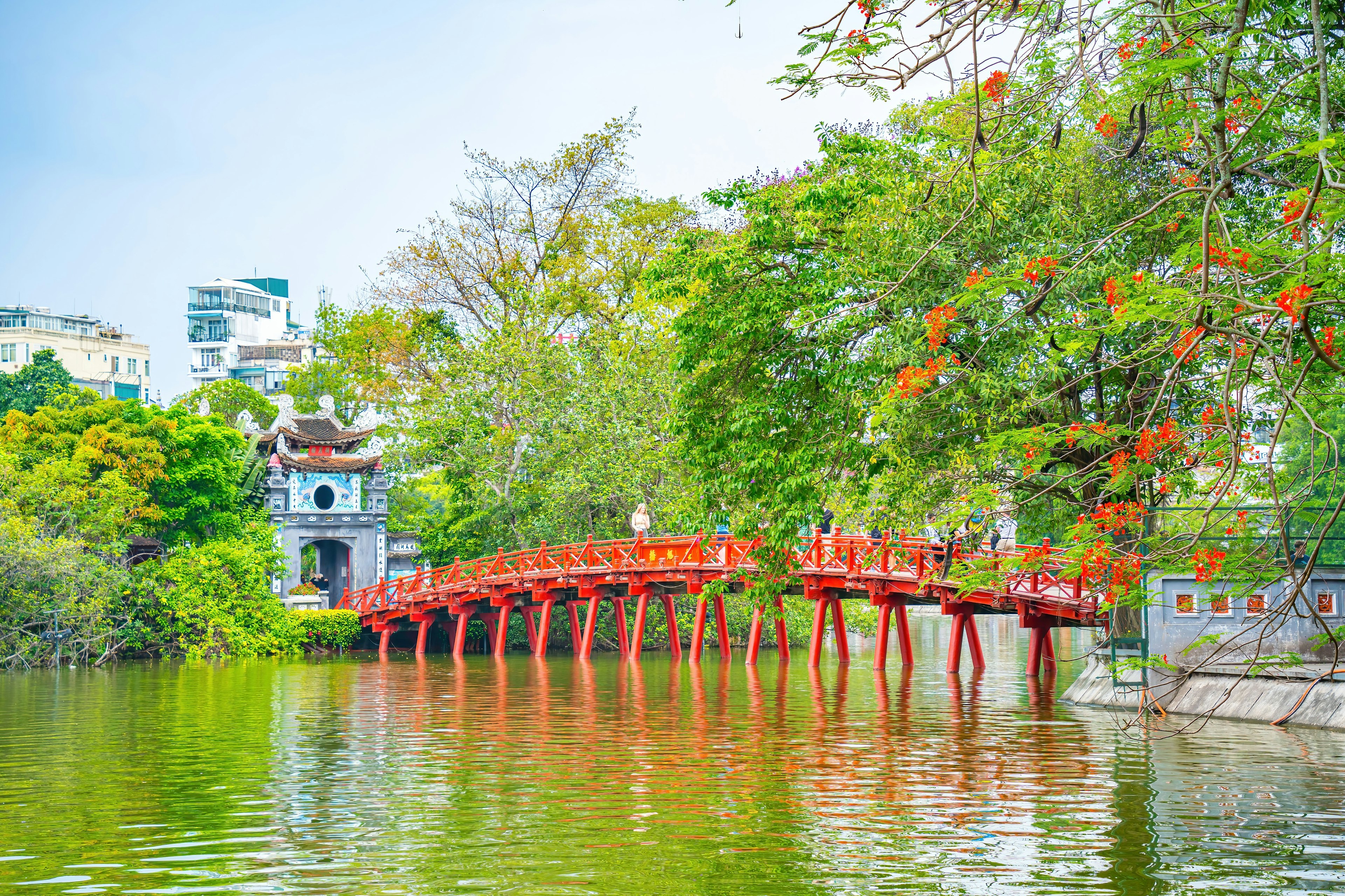 A red bridge spans a lake with bright green trees on its shore
