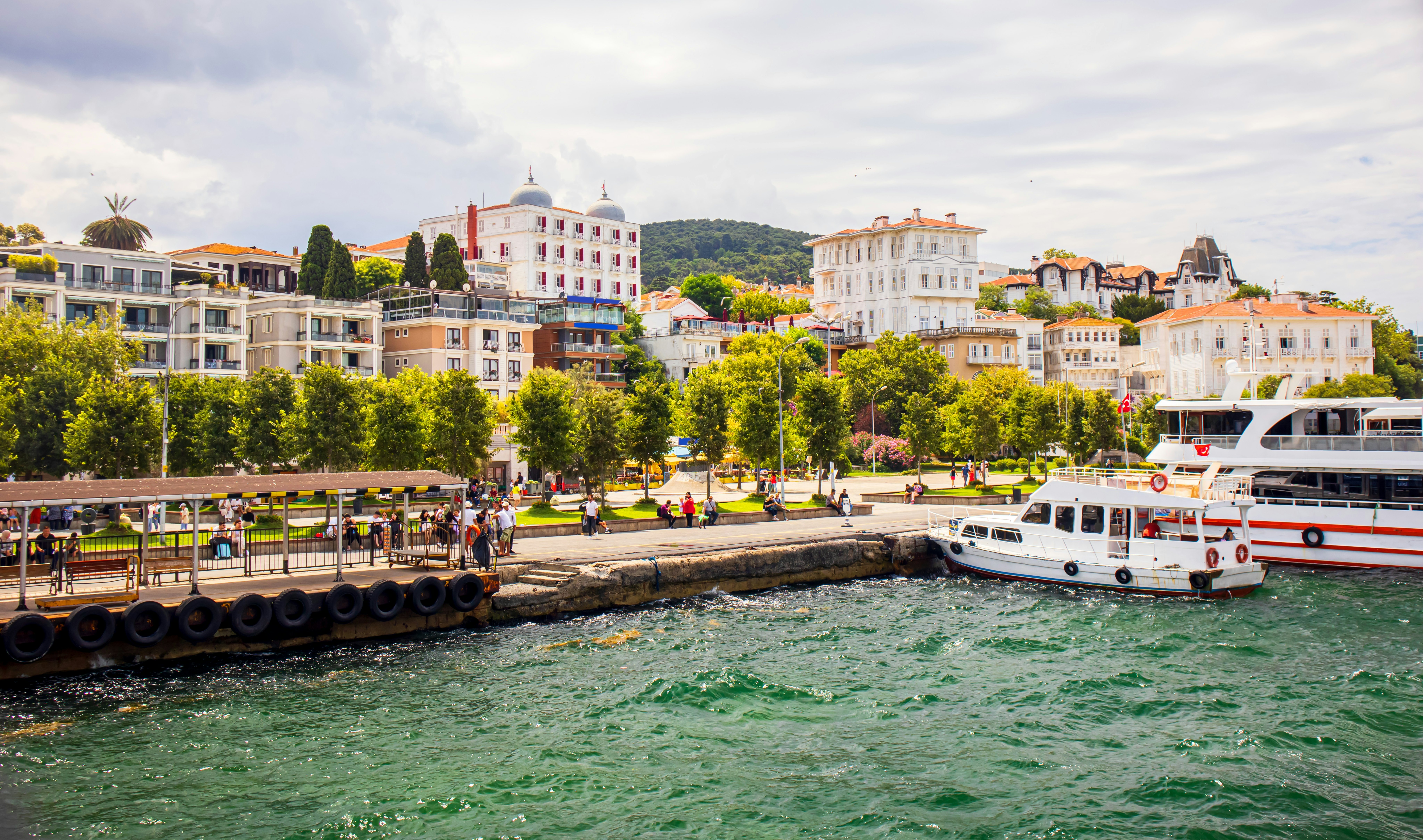 Two ferryboats dock at the waterfront of an island. A row of trees and elegant apartment buildings are seen near the water.