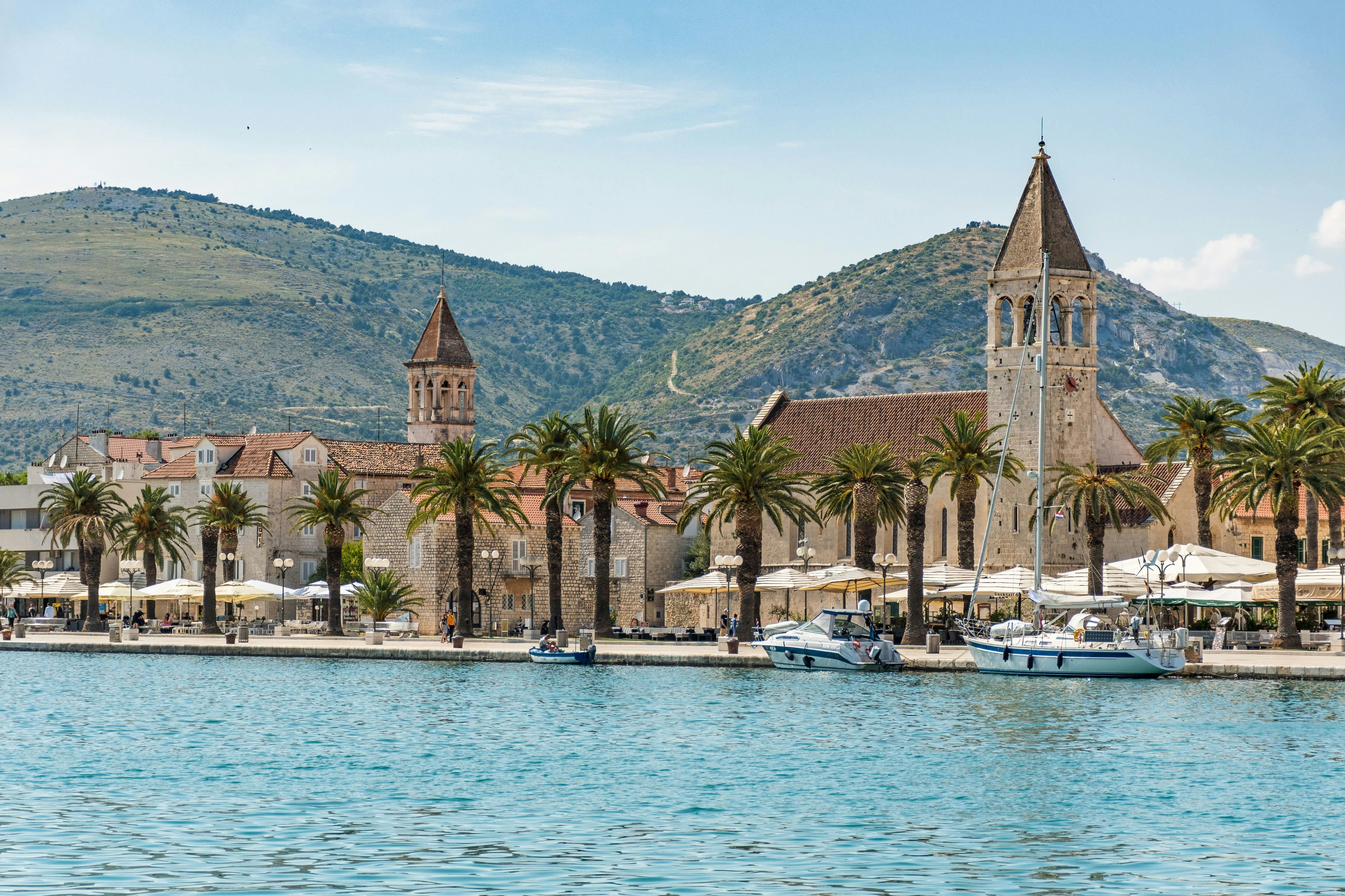 A palm-tree lined waterfront with three boats moored in the calm waters. Two church towers stand tall nearby.
