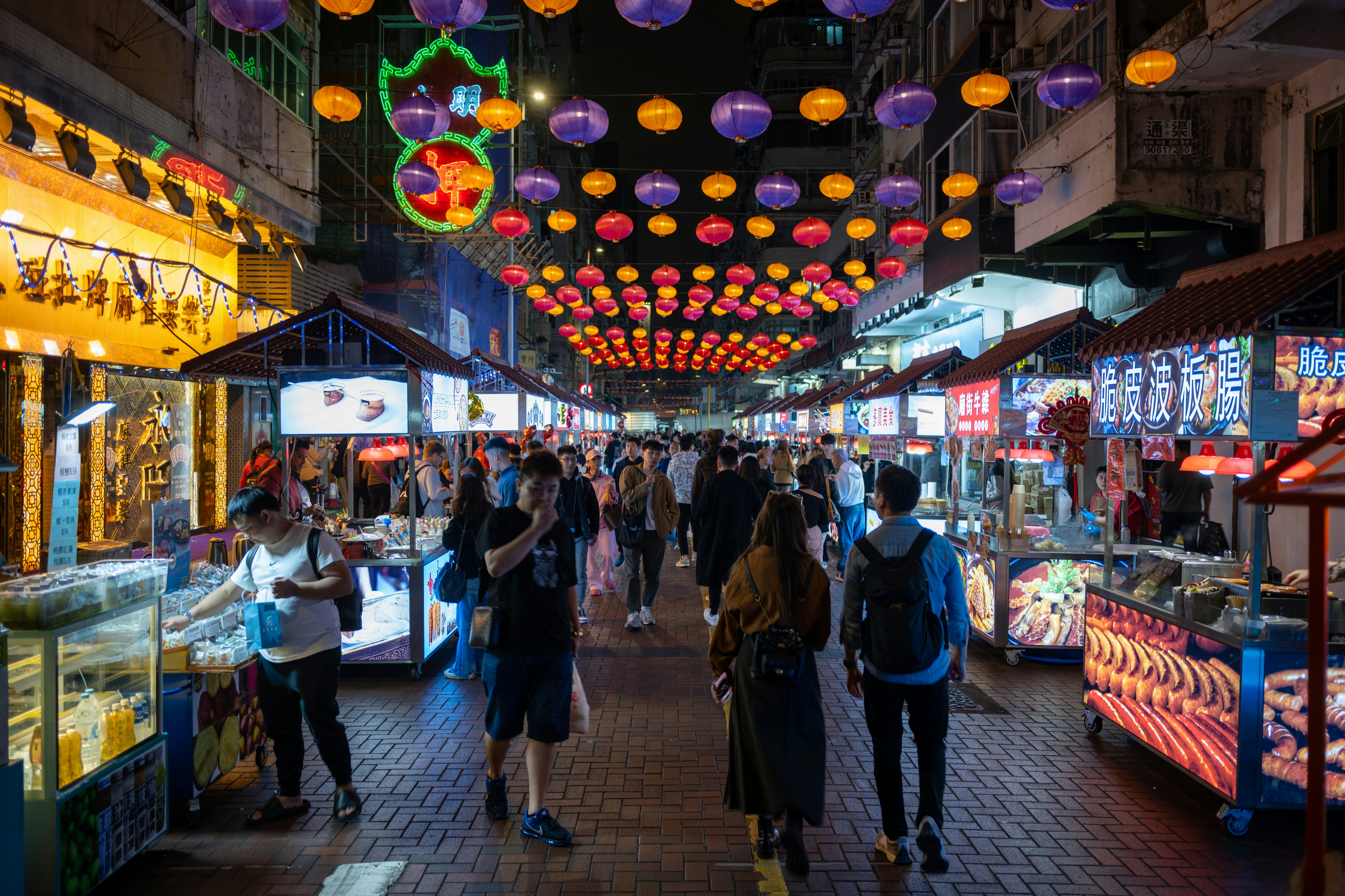 People walk past food stands at a night market, with multicolored lights overhead.
