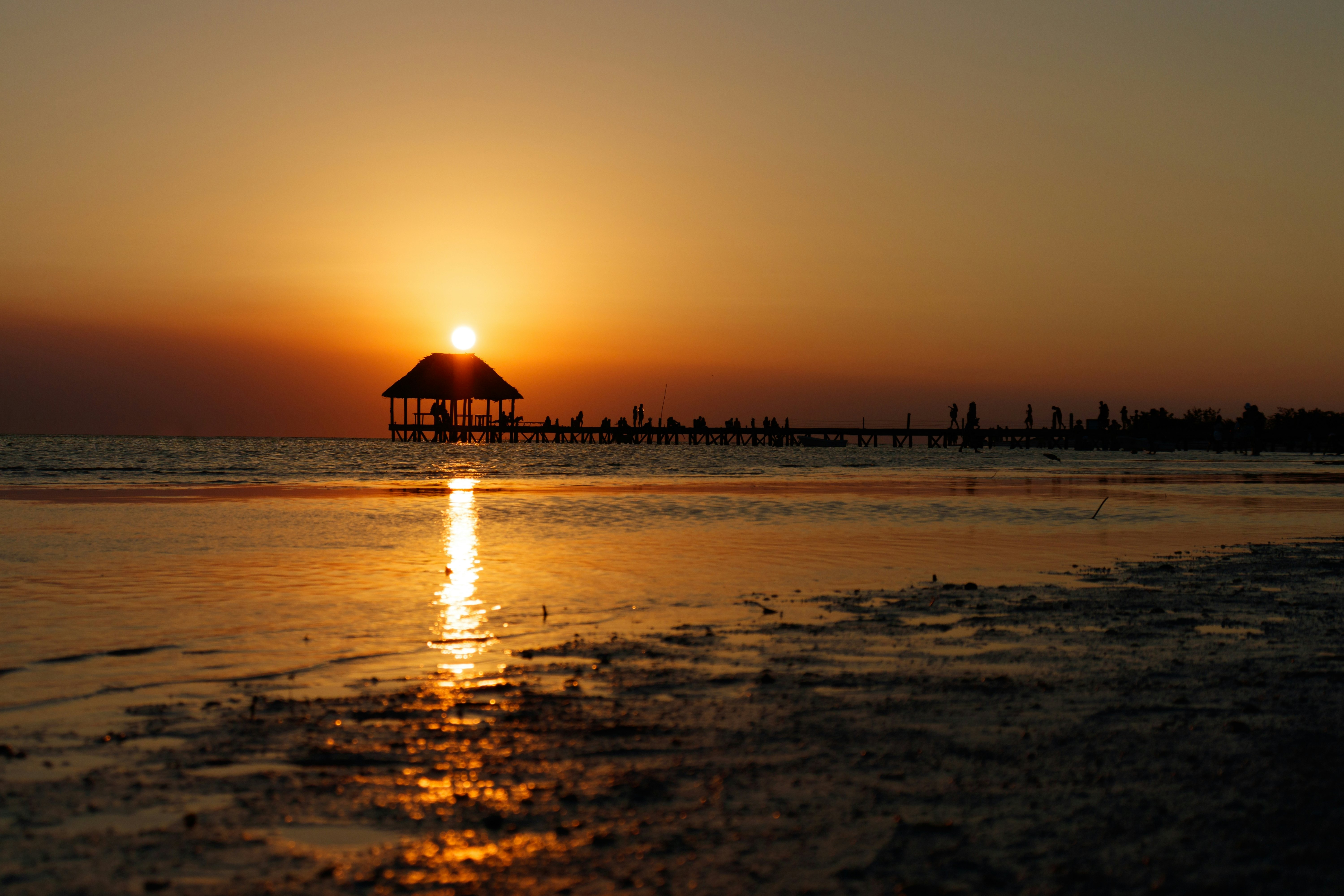 A silhouette of a pavilion at the end of a long pier extending from a beach at sunset.
