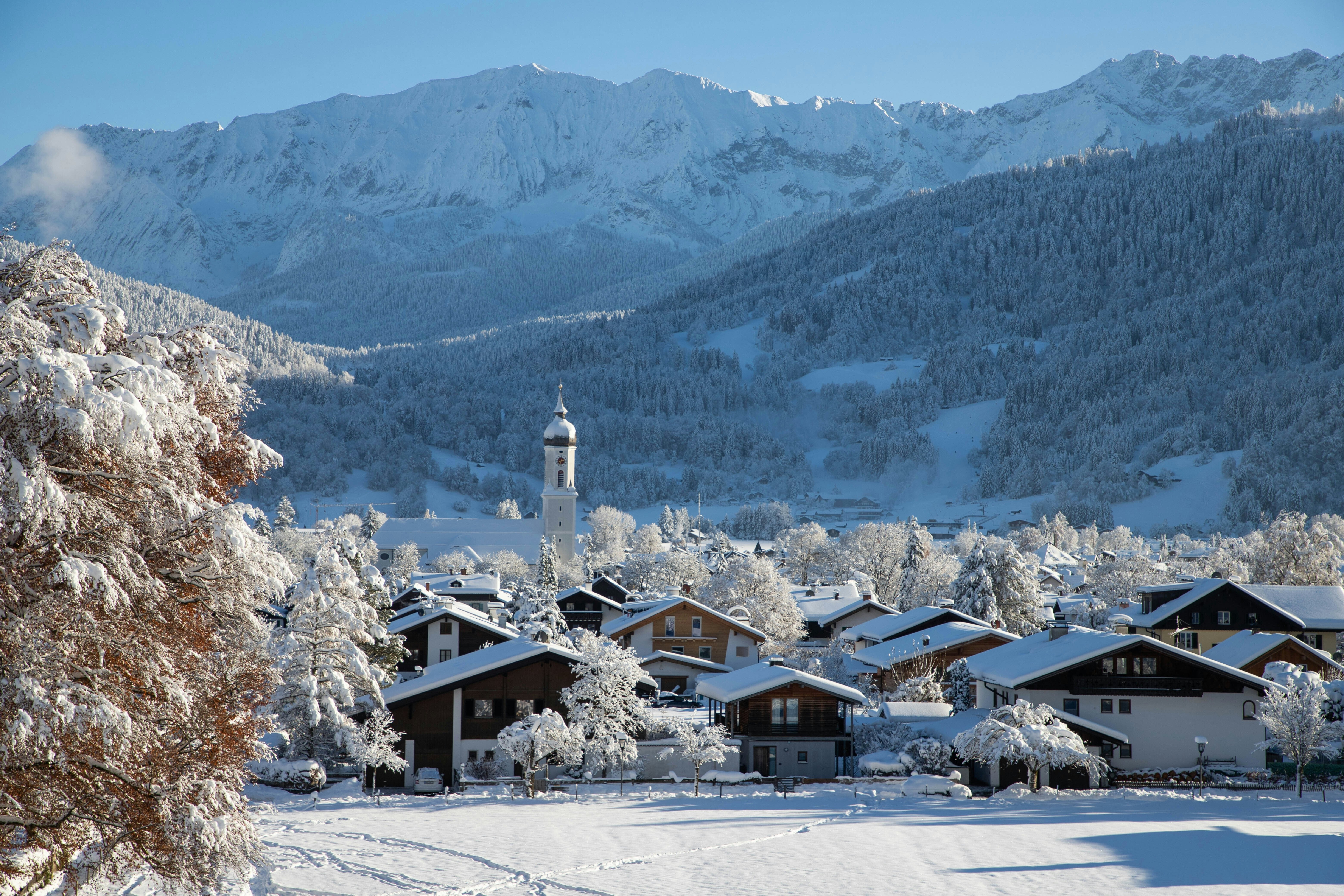 A view of a village: houses have low-angled roofs and a church spire towers above them; everything is covered in snow. Snowy mountains rise in the background.