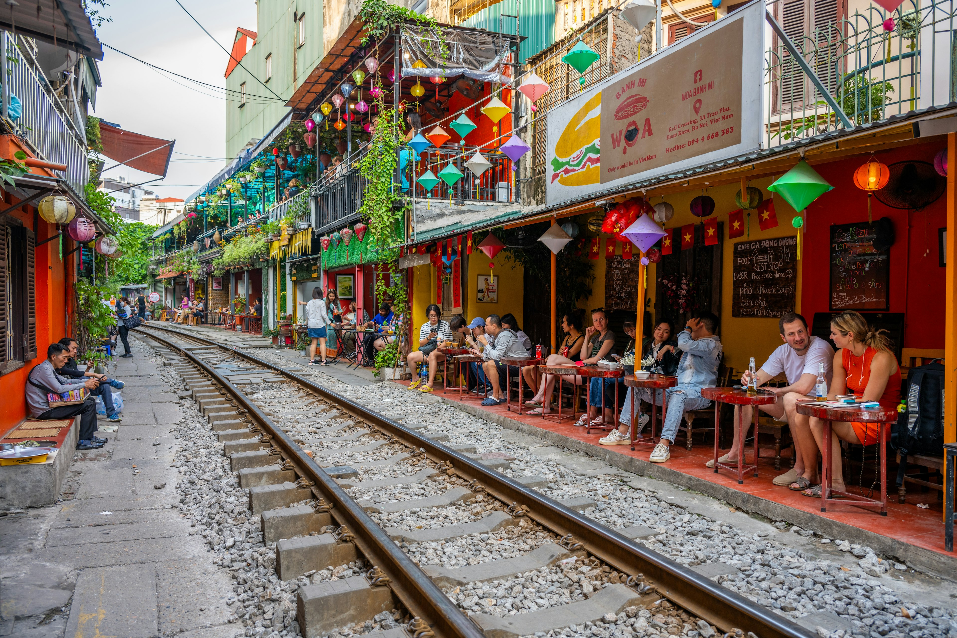 People dine outside of restaurants that line a narrow train alley. The restaurants have colorful decorations hanging.