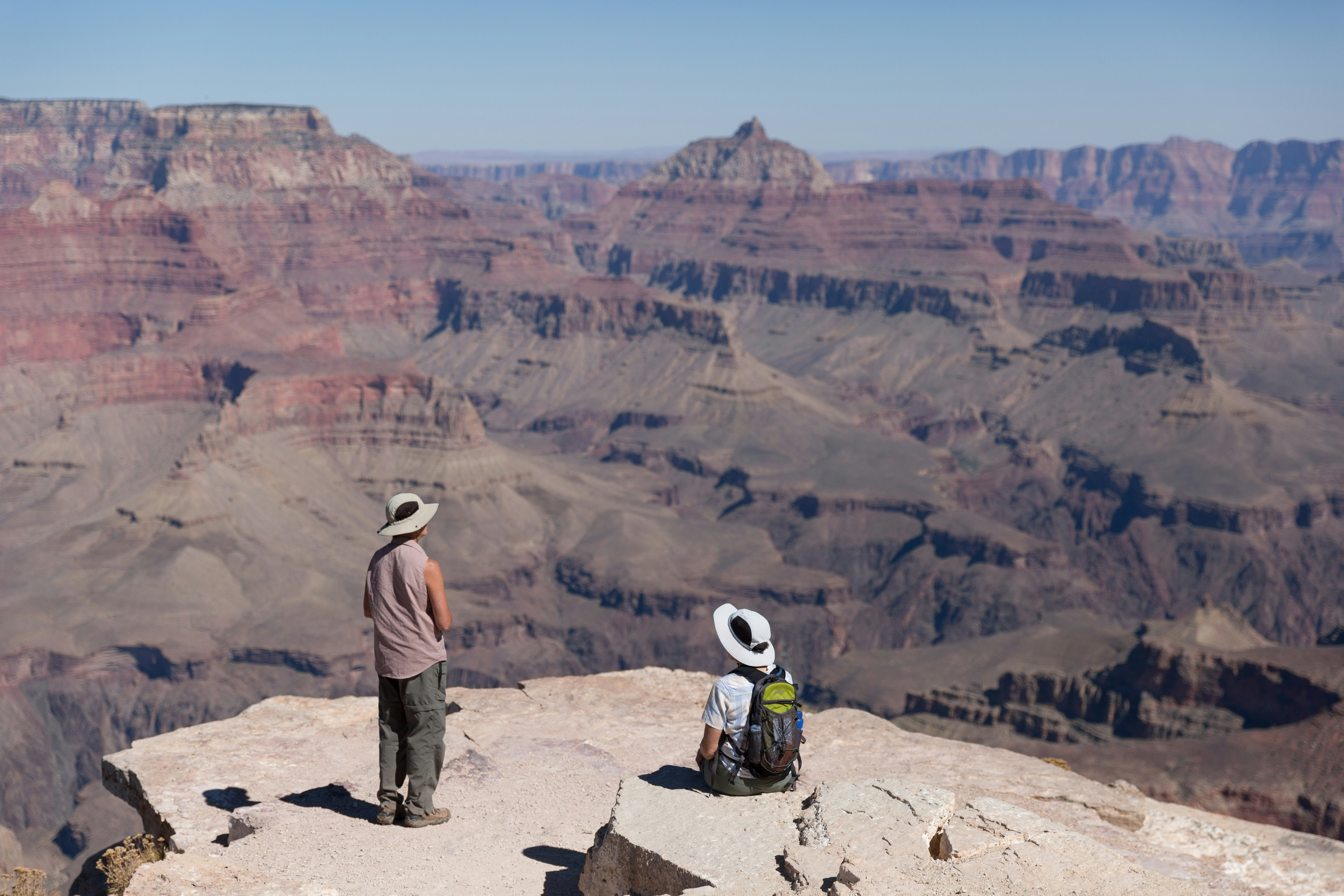 One person standing and another sitting, contemplating the wide landscape of Grand Canyon at Shoshone Point.