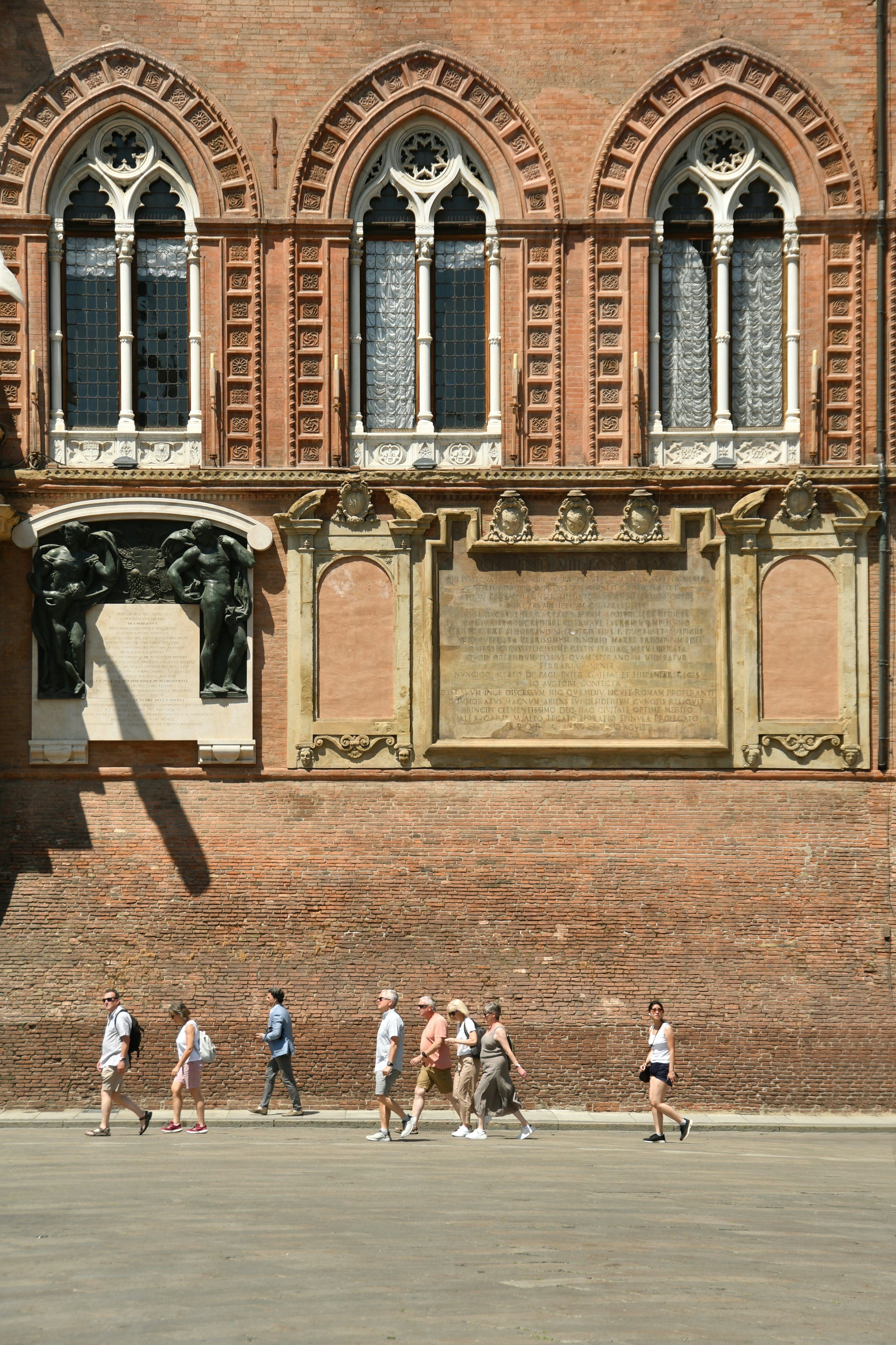 People walk through a city square on a sunny day passing a large red-brick building with statues and engravings in its exterior walls.