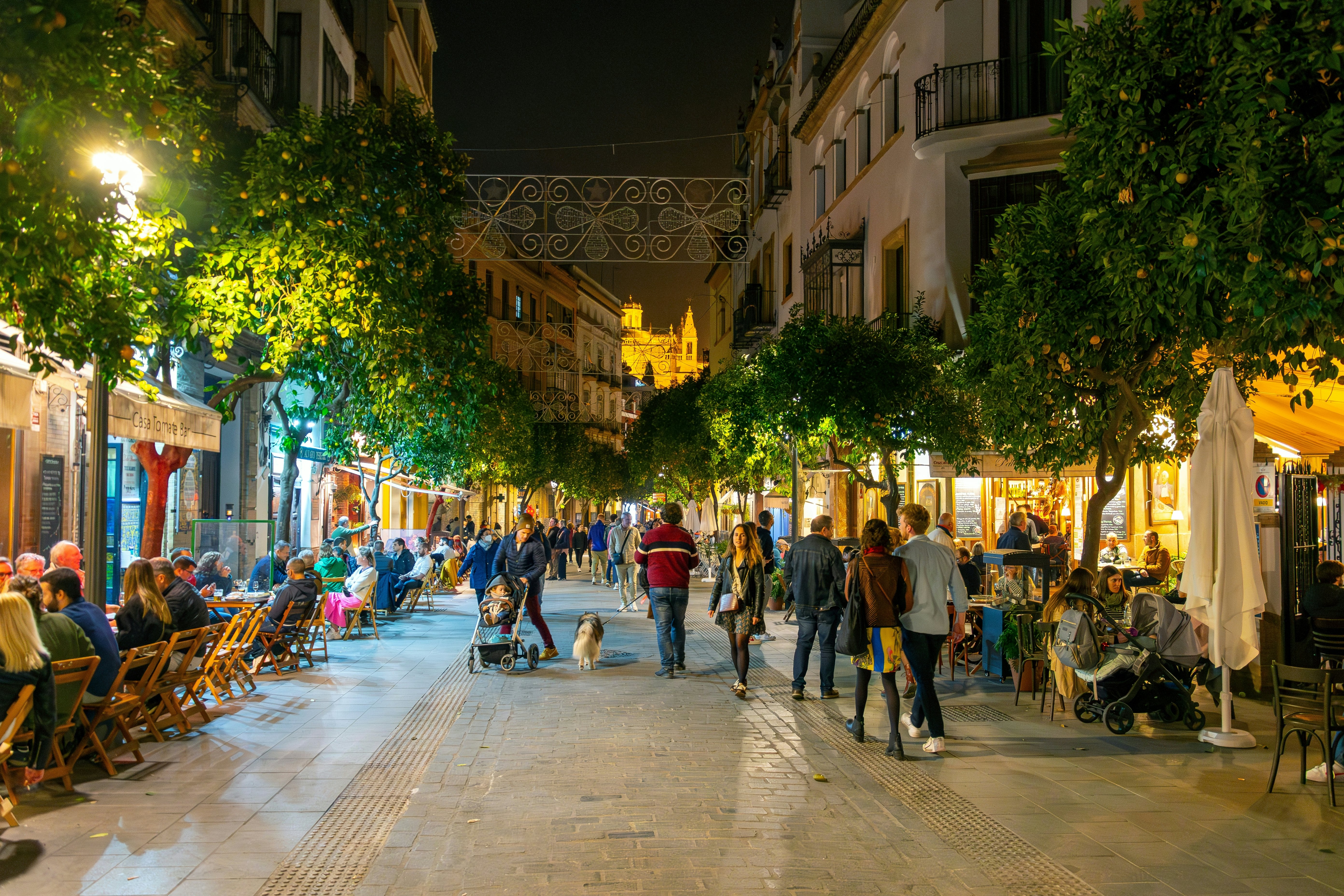 Seville, Spain: The lights of the Seville Cathedral in the distance from a busy touristic street of sidewalk cafes in the Barrio Santa Cruz district of central Seville, Spain.