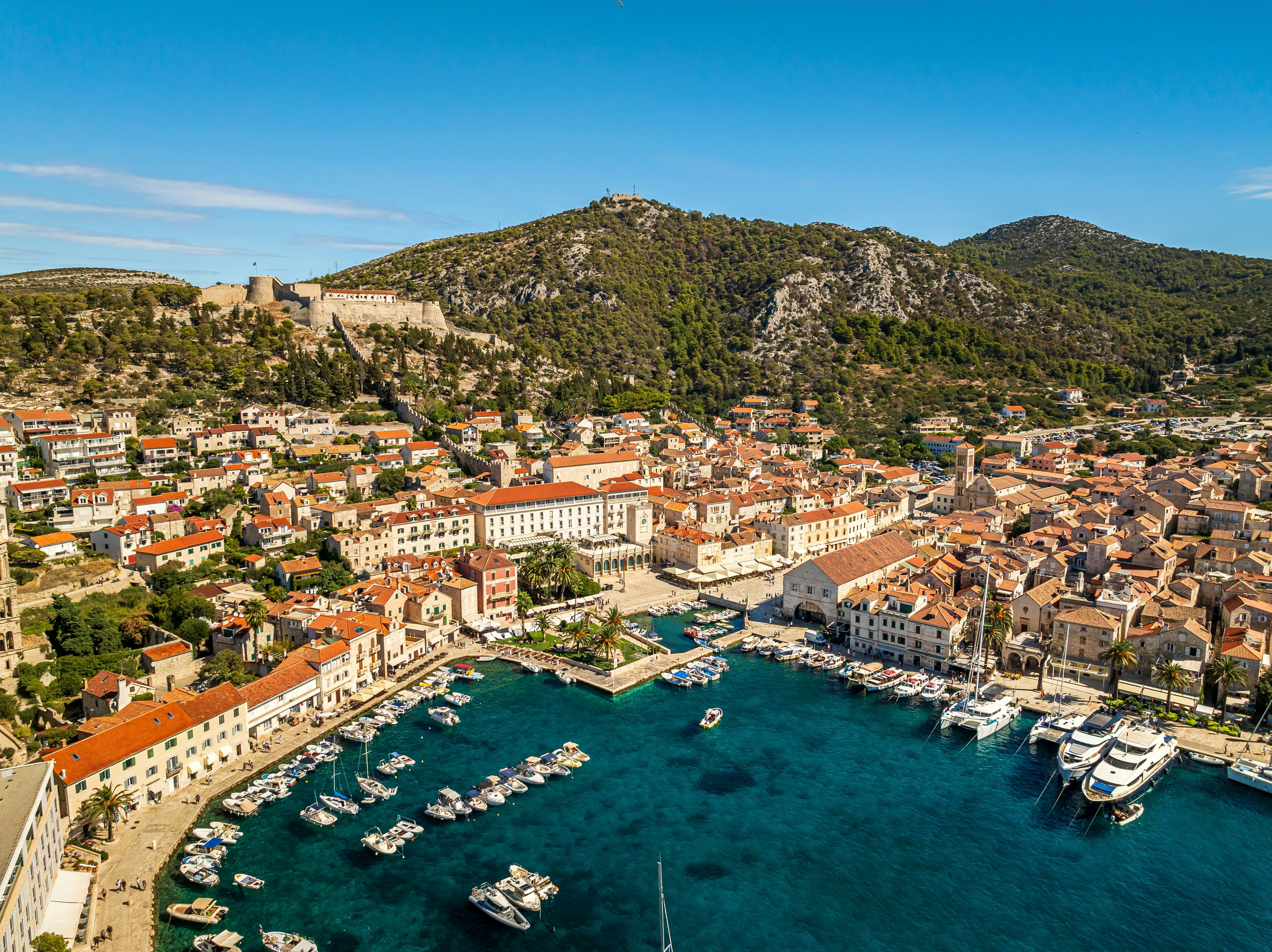A Mediterranean town with boats docked in its a harbor, red-roofed buildings and a fortress high on the hill above.