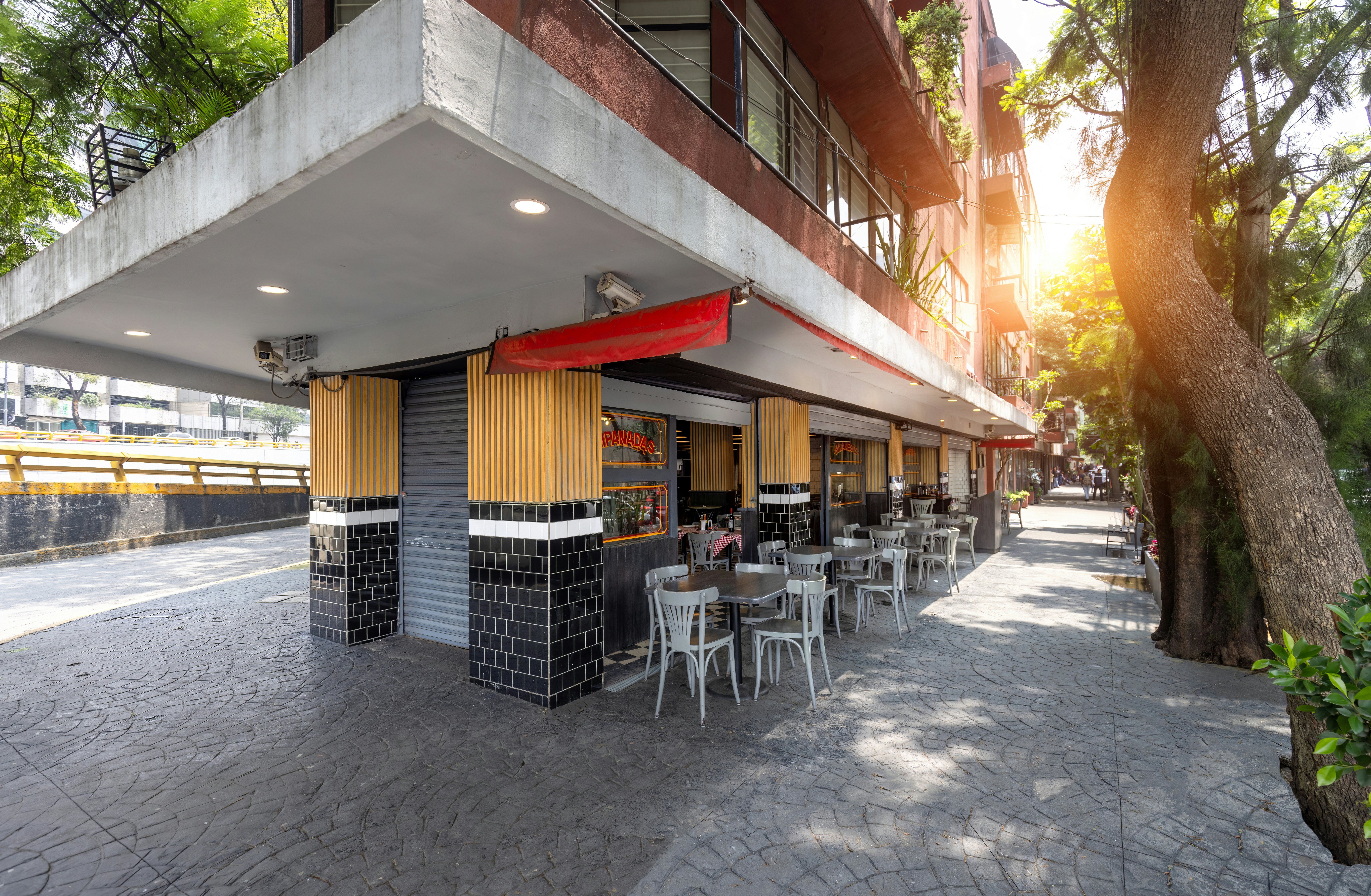A view of a restaurant on a narrow street corner. The sunset light shines on a sidewalk lined by large trees.