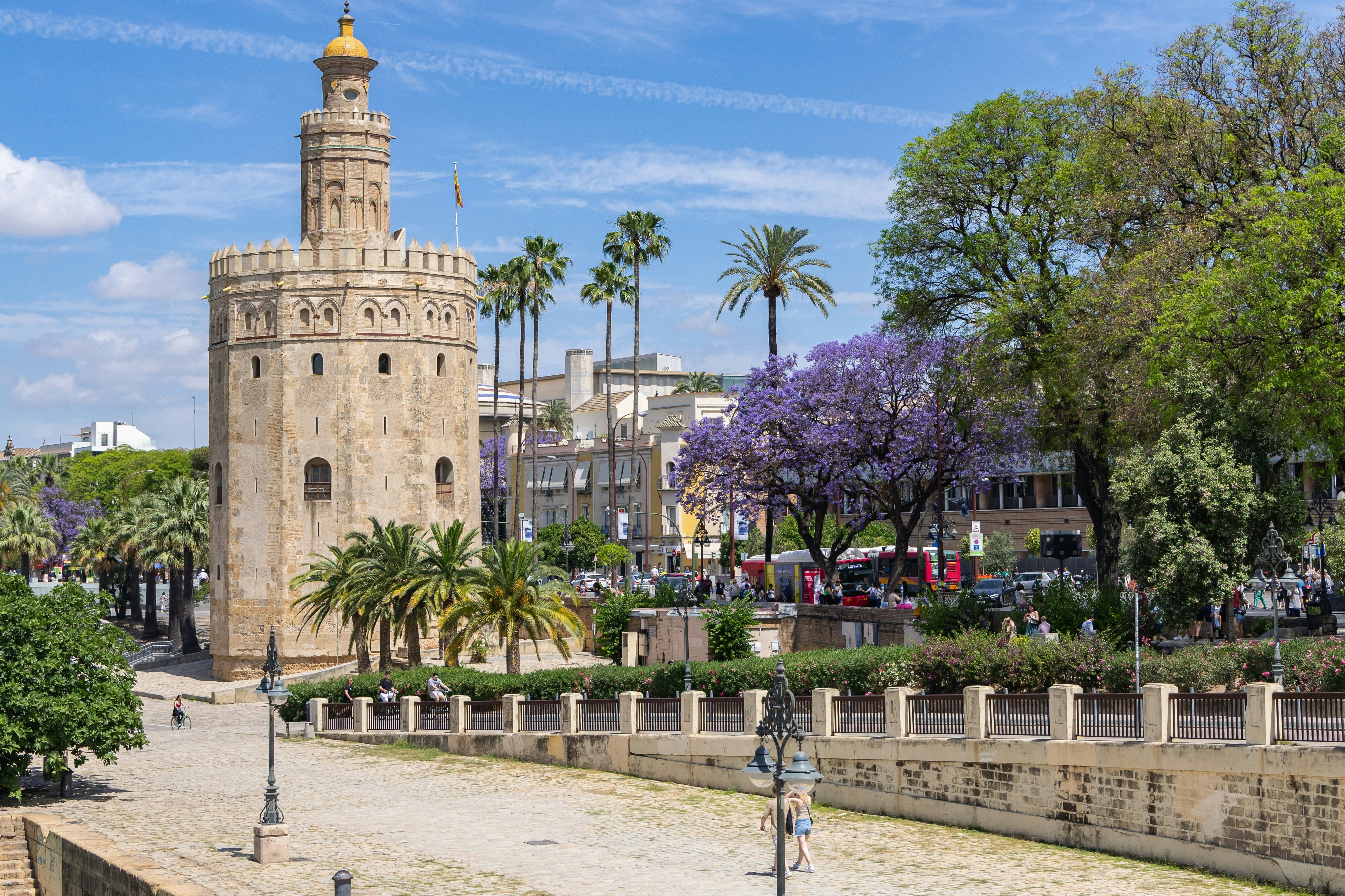 A stone tower stands along a wide sidewalk in a city. Trees bloom along the pathway.