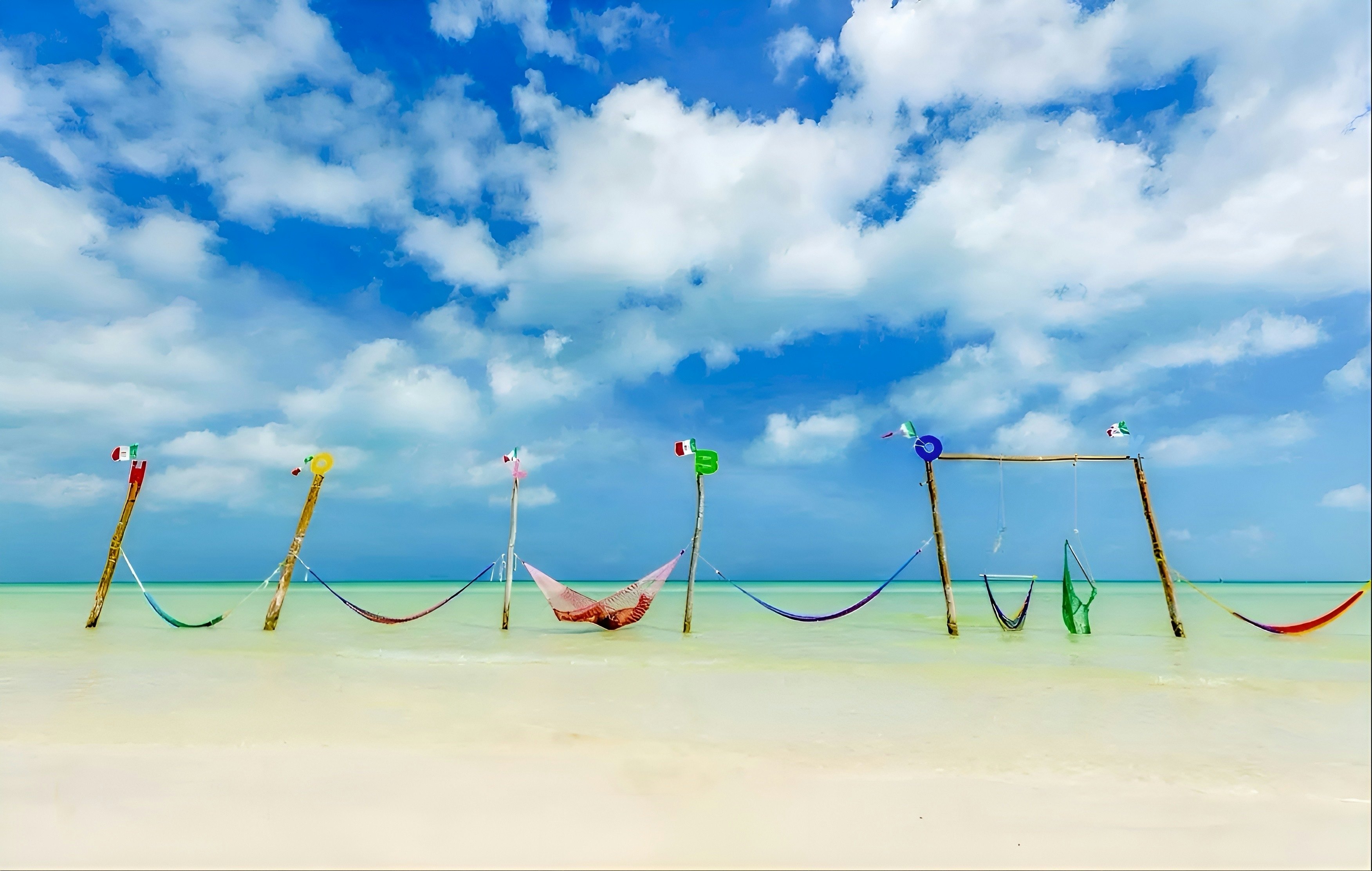 A line of seven multicolored hammocks attached to posts set in shallow turquoise water.