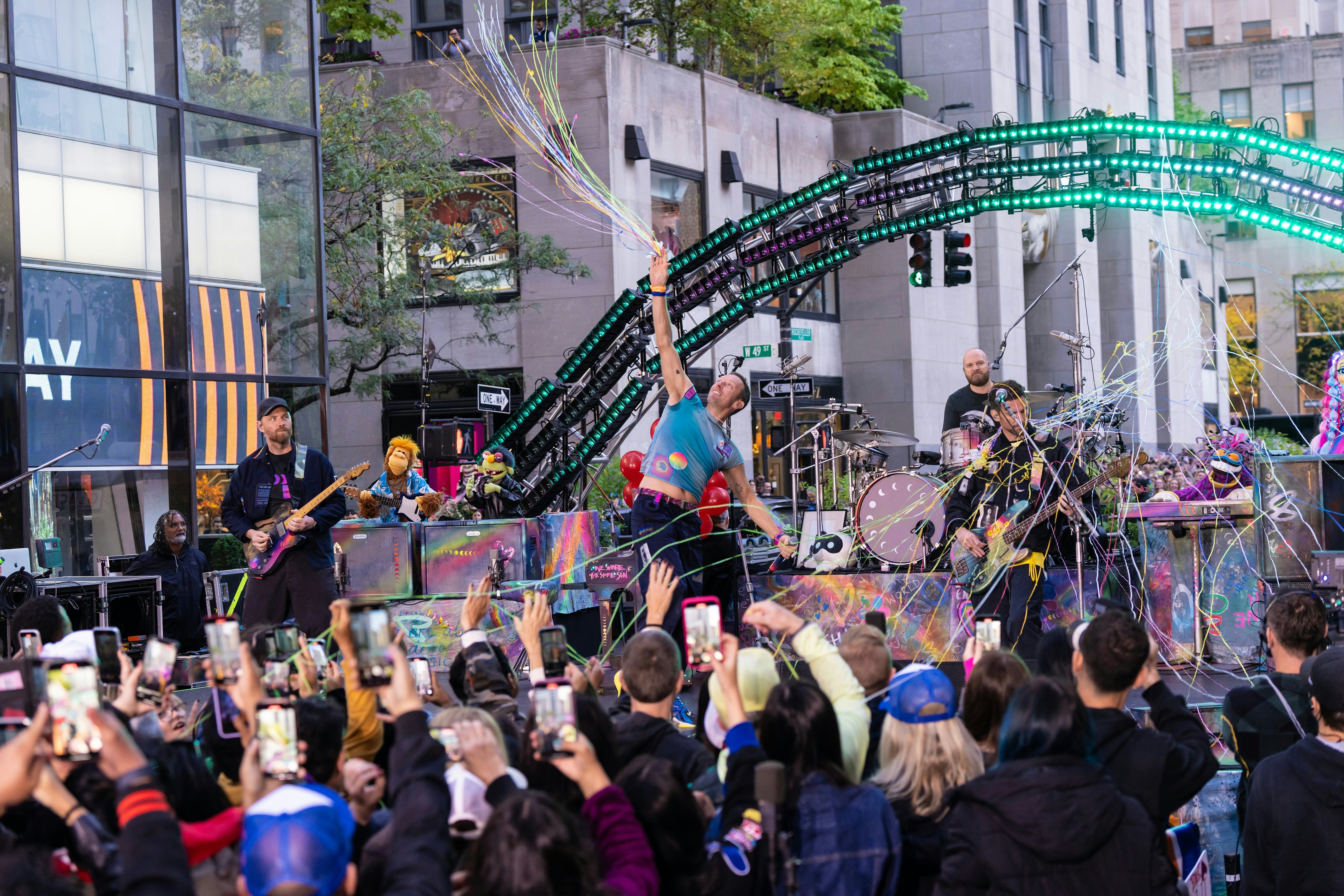 A band performs in front of a live audience during a taping in a city plaza.