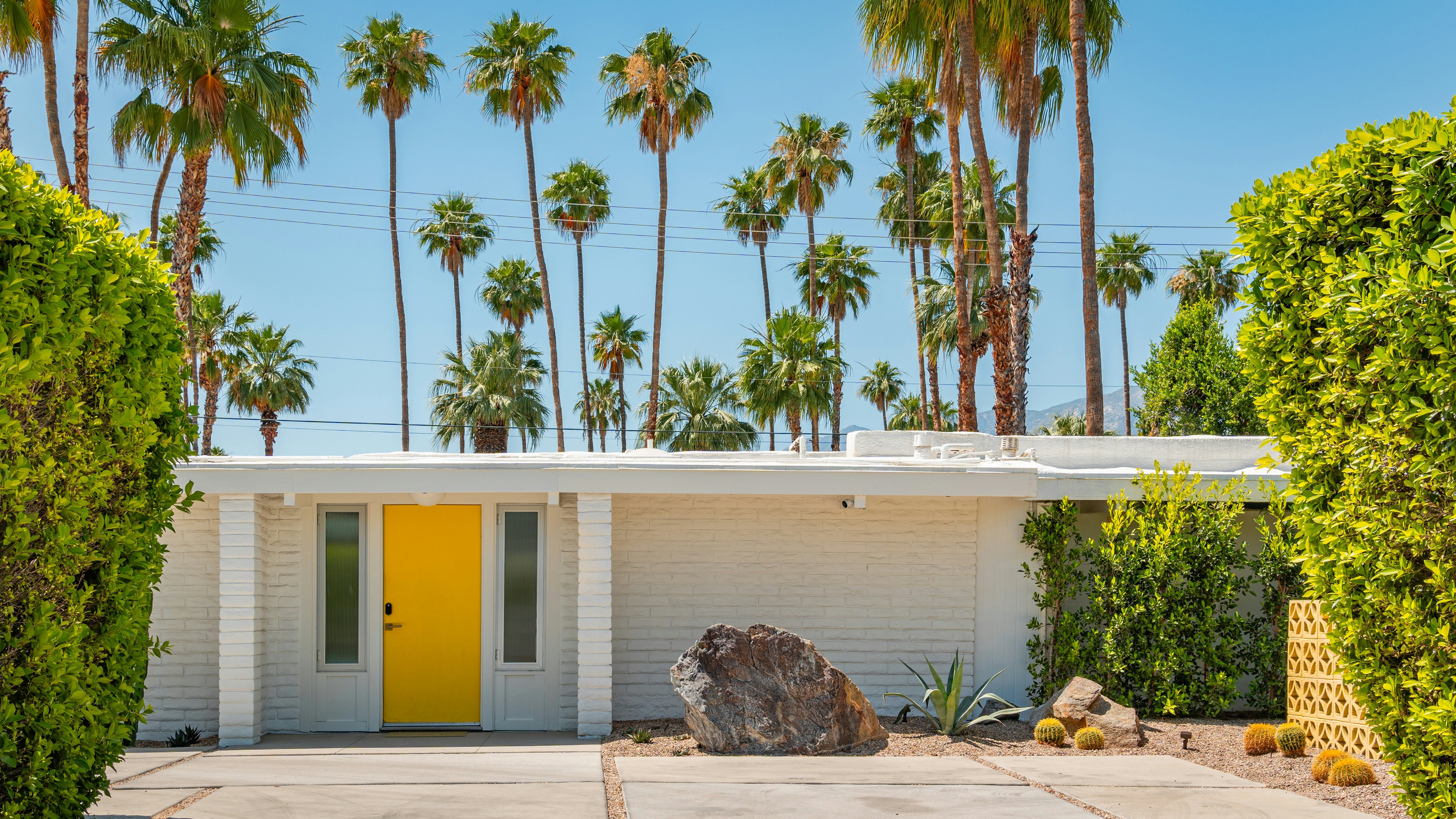 A low-rise property with a simple design of straight lines and a yellow front door. Palm trees tower over head.