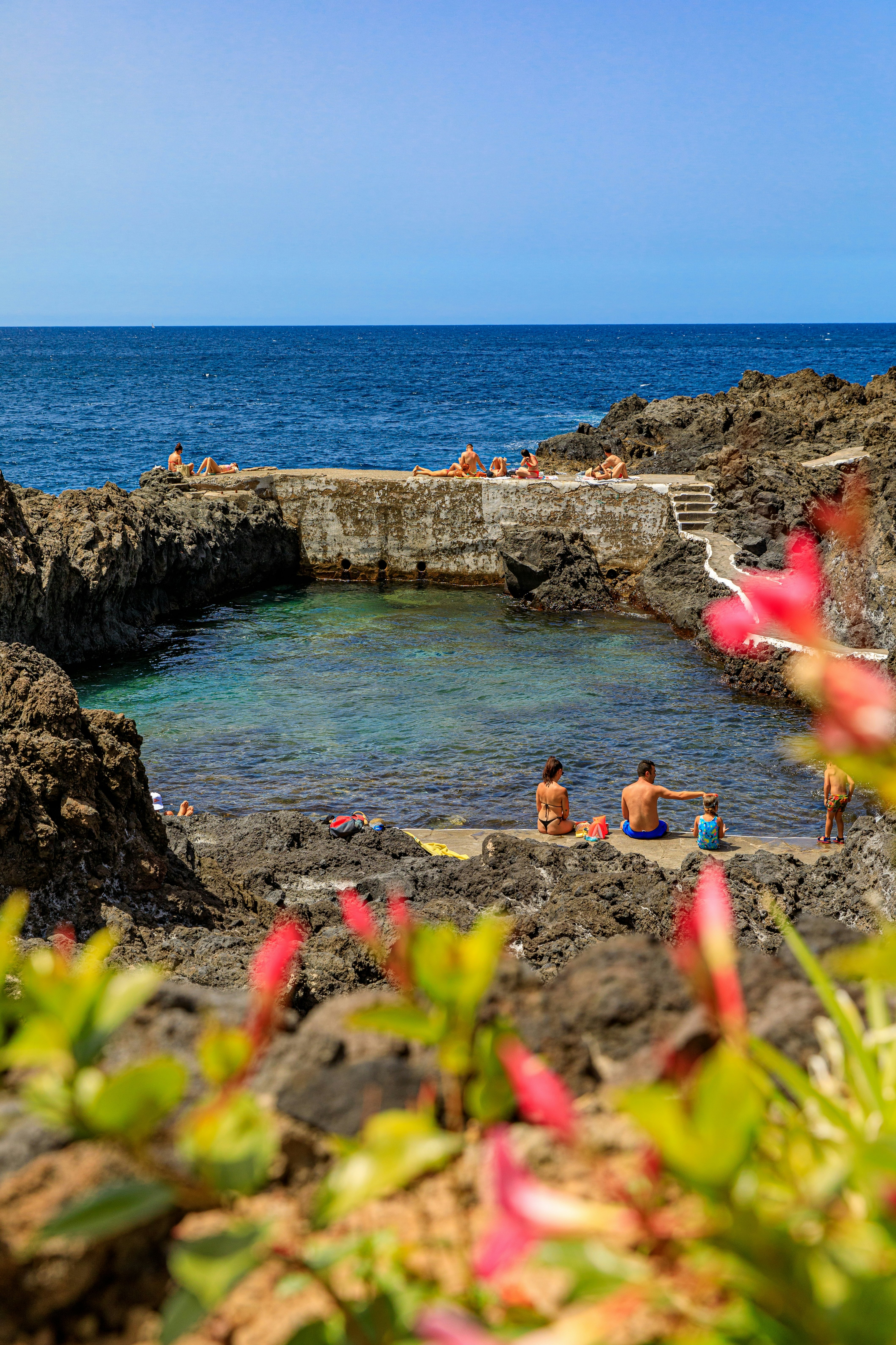 People sitting around the wall of a natural pool in the sea. Pink flowers are in the foreground.