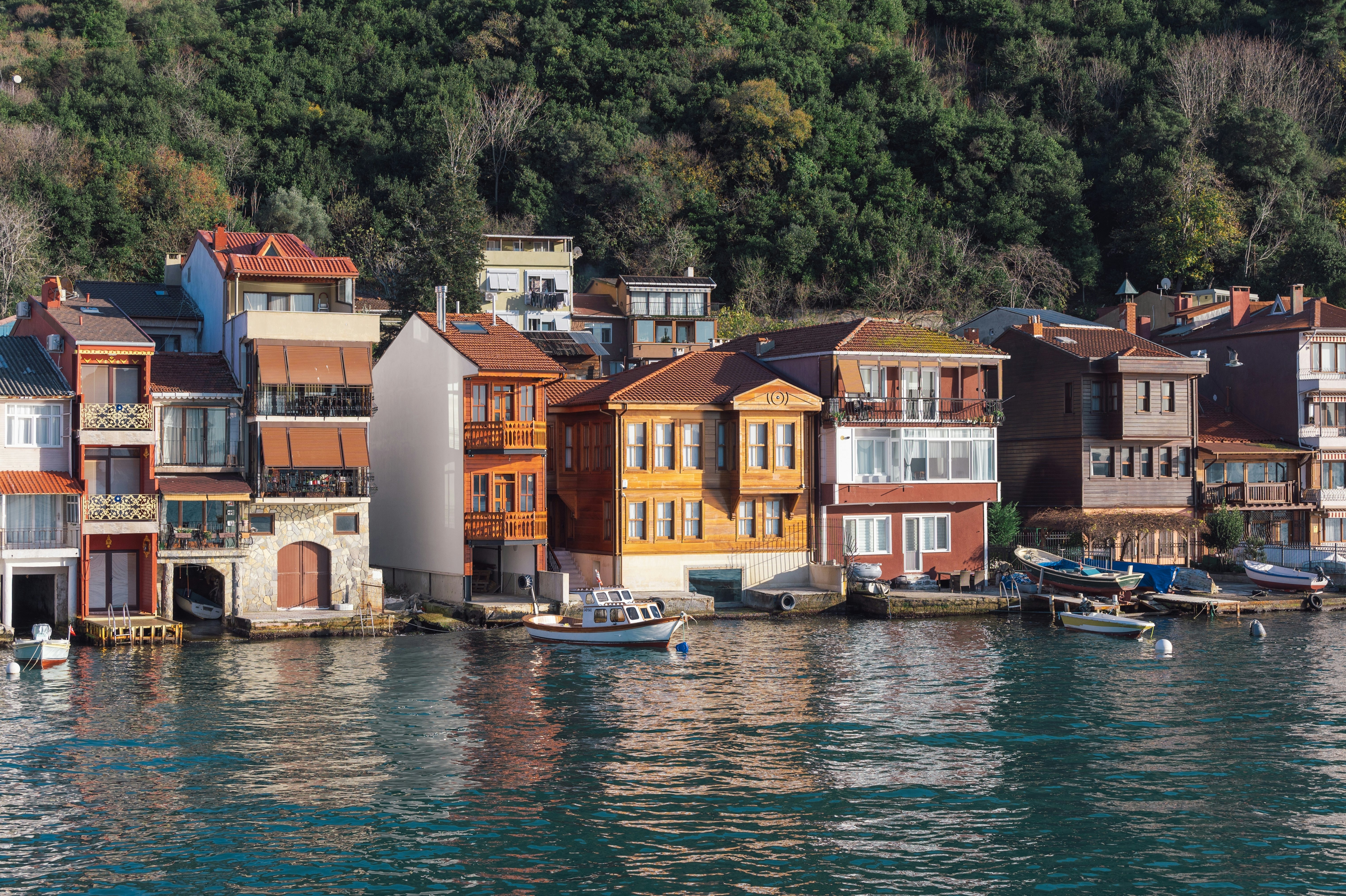 Water laps a row of four-story houses, with boots moored in front and a hilly forest behind.