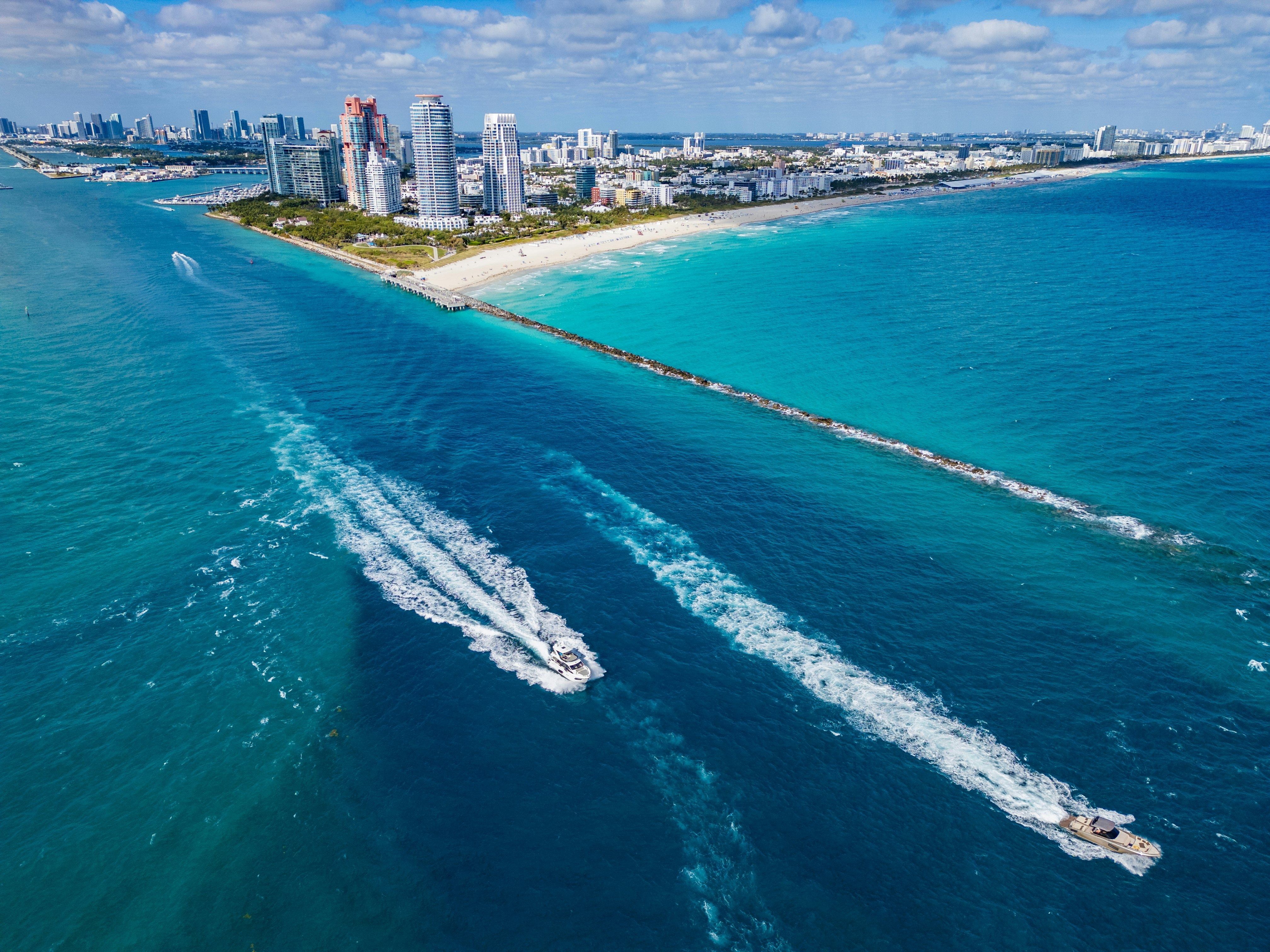Two boats leave a long wake in blue water; a city skyline is in the background.