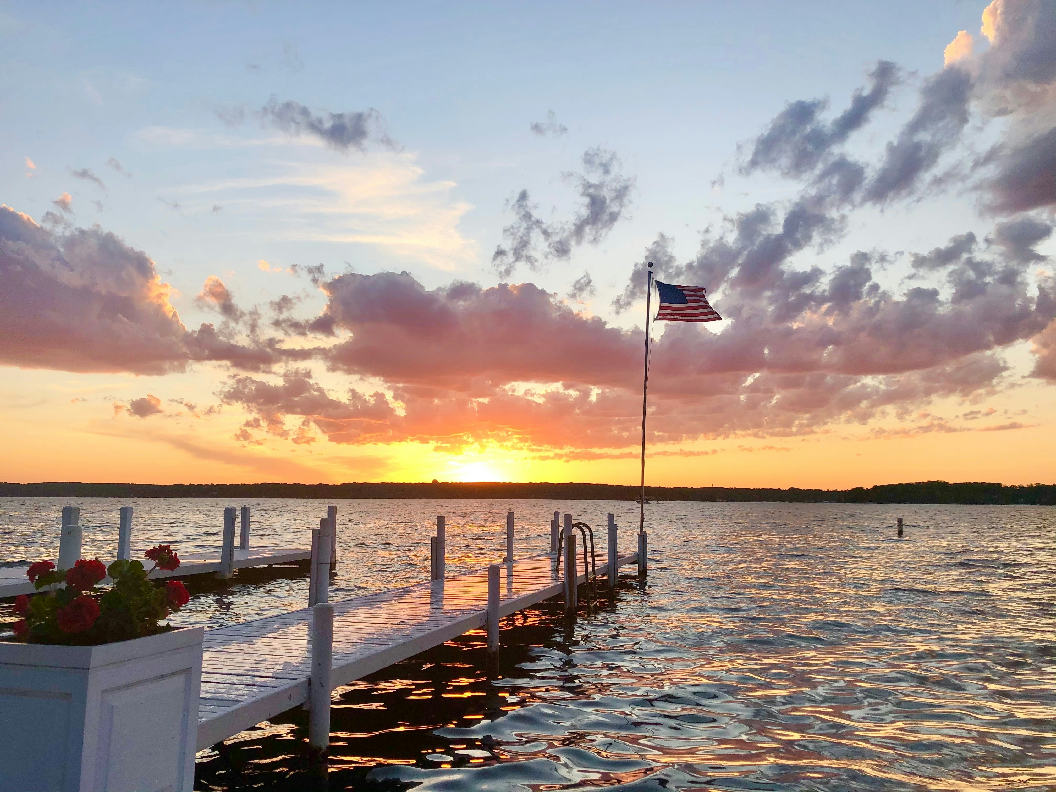 A an American flag flies from a pole at the end of a pier in a lake at sunset.