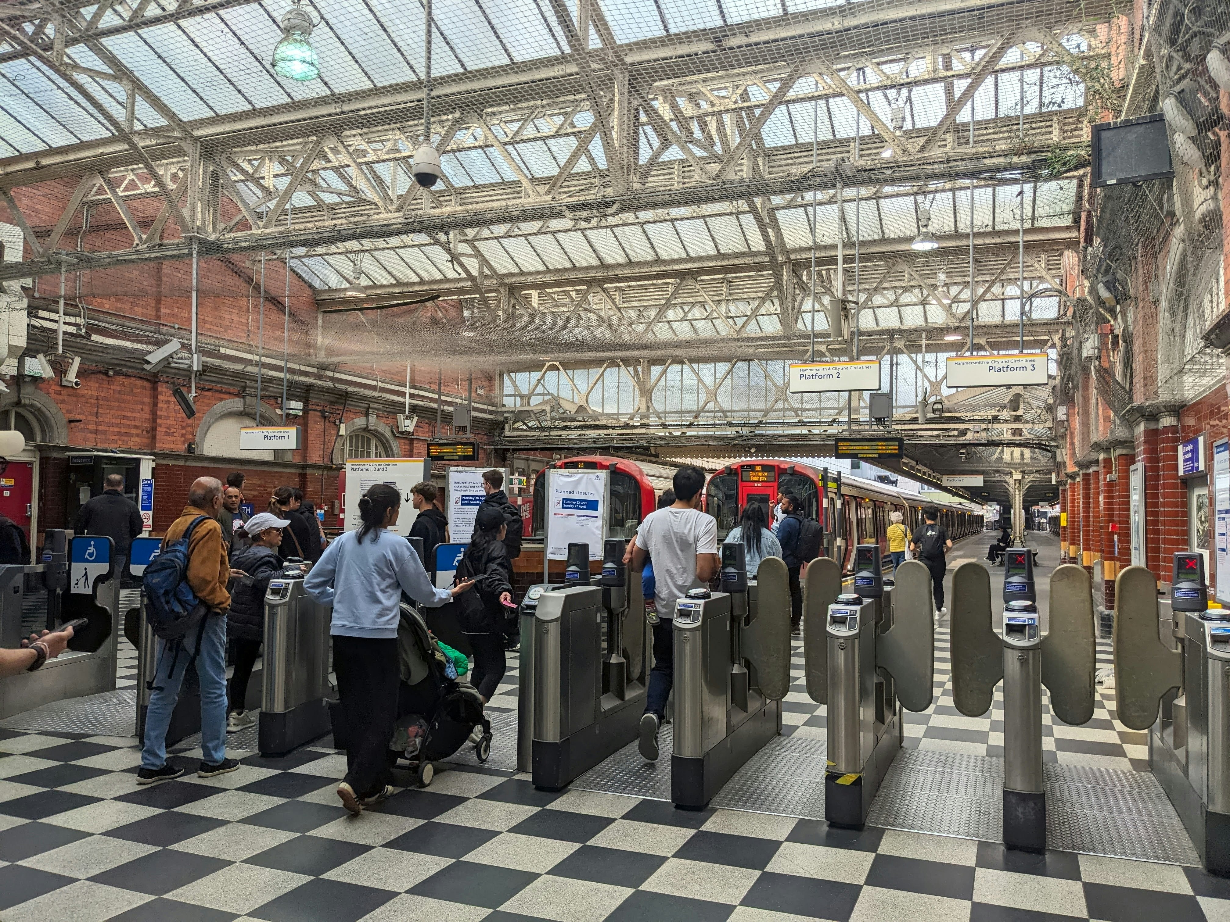 People pass through ticket barriers leading into a metro station in a big city. Checkerboard tiles are on the floor, while skylights flood the station with light.
