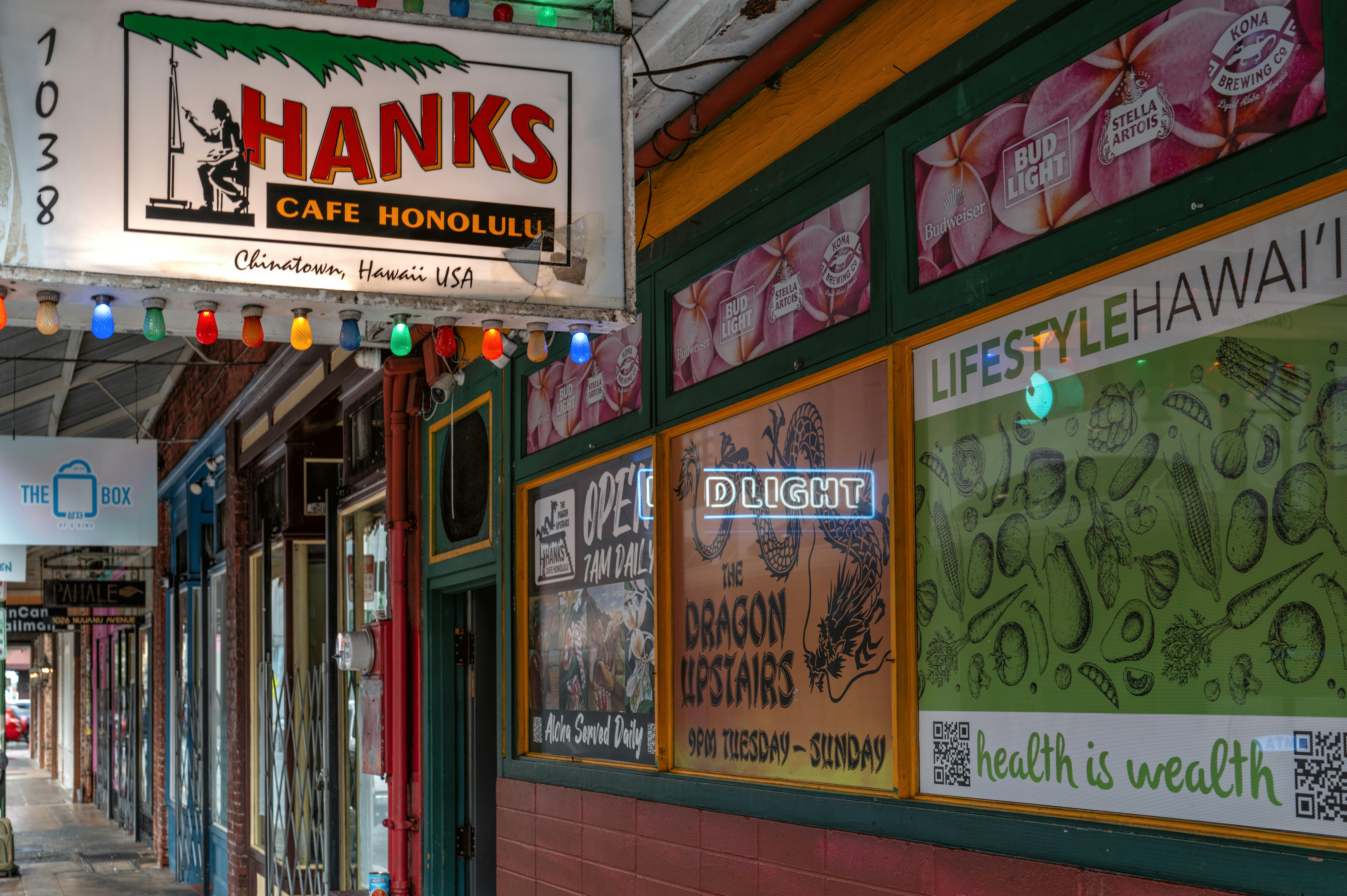 View of a block of colorful storefronts. A lighted sign reads "Hanks Cafe Honolulu, Chinatown, Hawaii, USA." A poster on a window advertises The Dragon Upstairs.