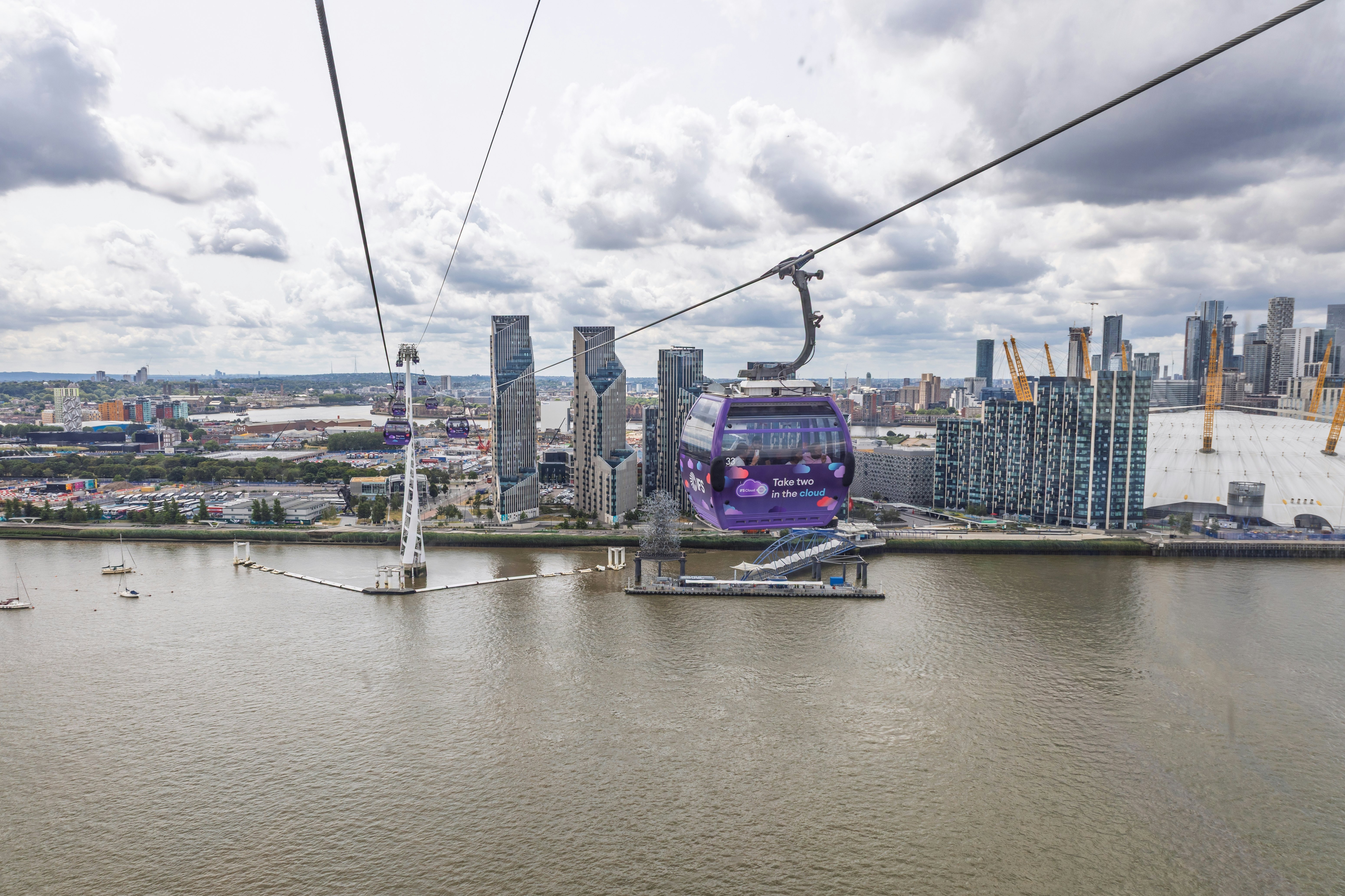 A purple gondola on a high cable line crosses a river, with views of towers and a domed arena in view on the shore.