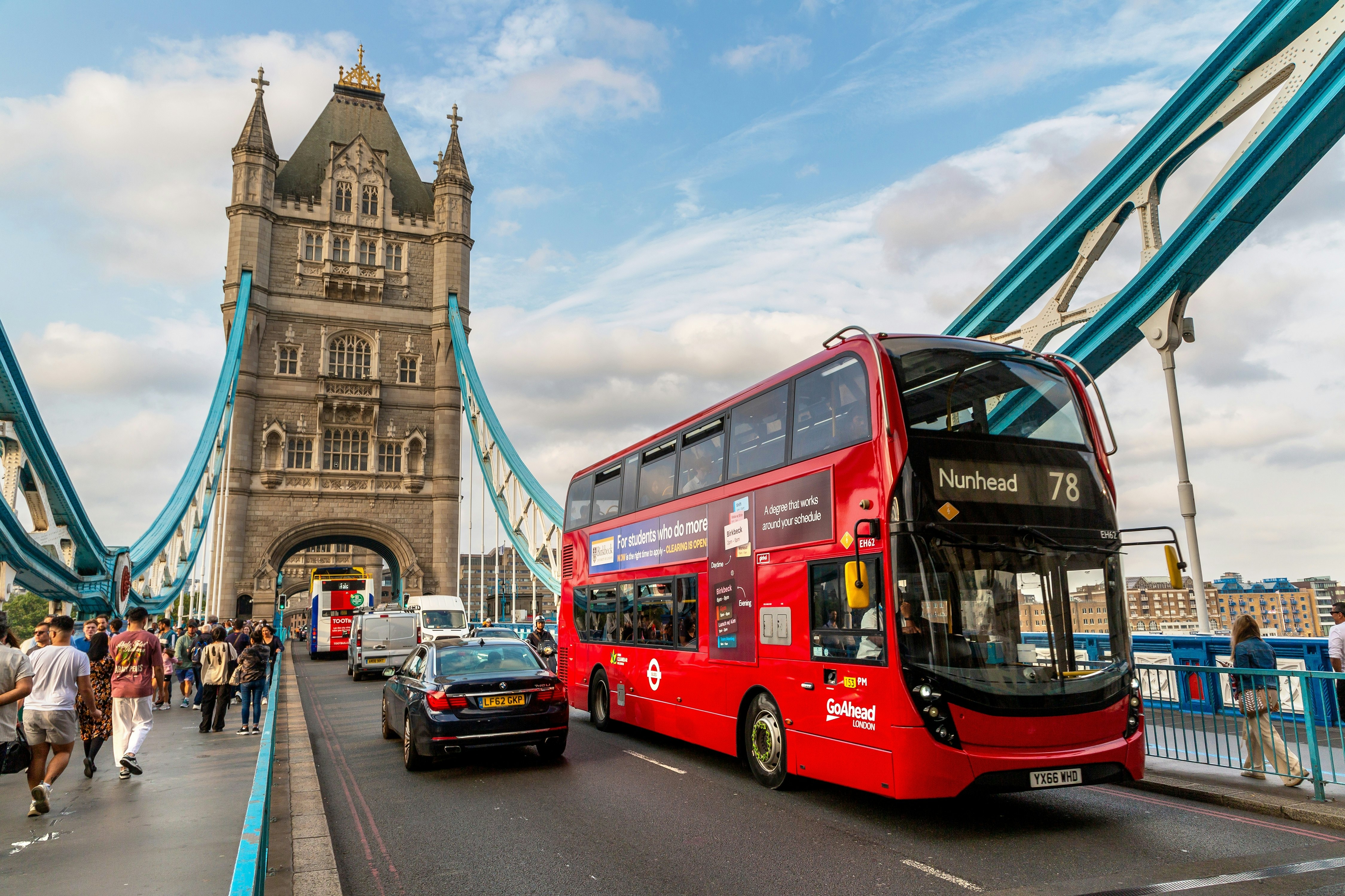A red double-decker bus drives alongside other vehicles across a bridge with blue cables. The elaborate tower of the bridge is seen behind the bus. People walk along footpaths on either side of the roadway.