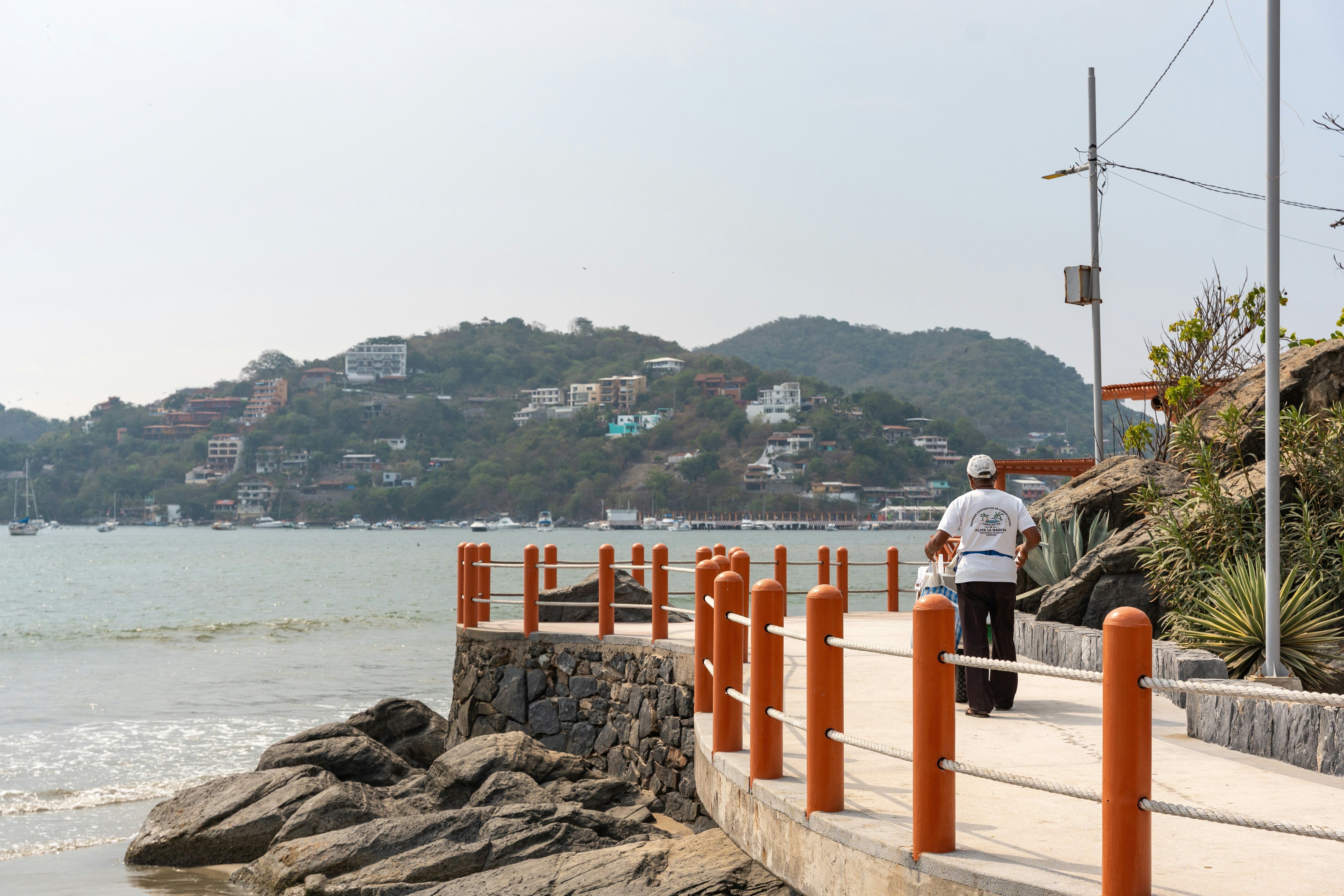 A man is seen from behind walking on a passage by the sea. A headland with houses and apartments is seen across the bay.