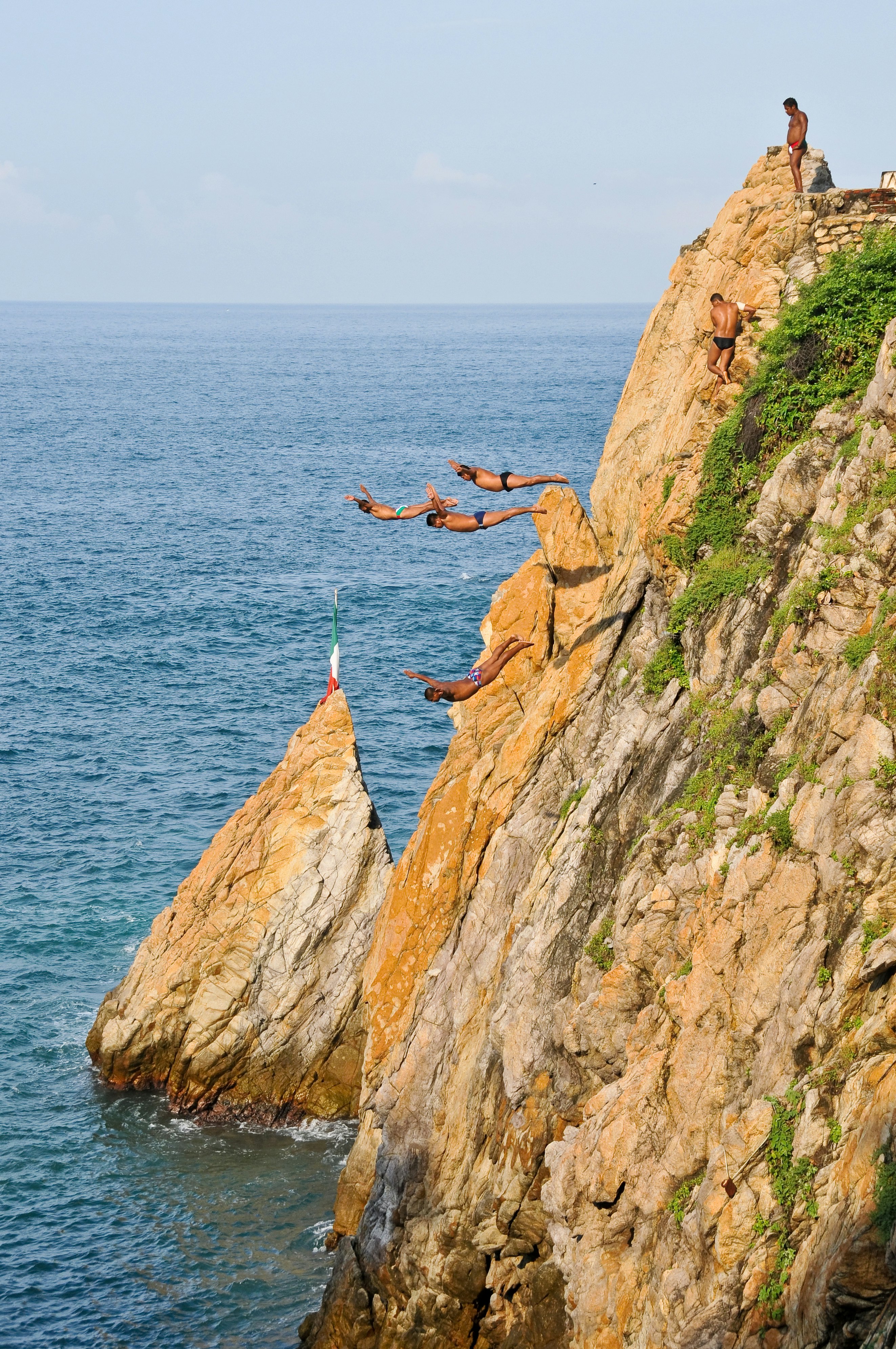 Four skilled cliff divers soar through the air to dive into the ocean on a rocky shore.