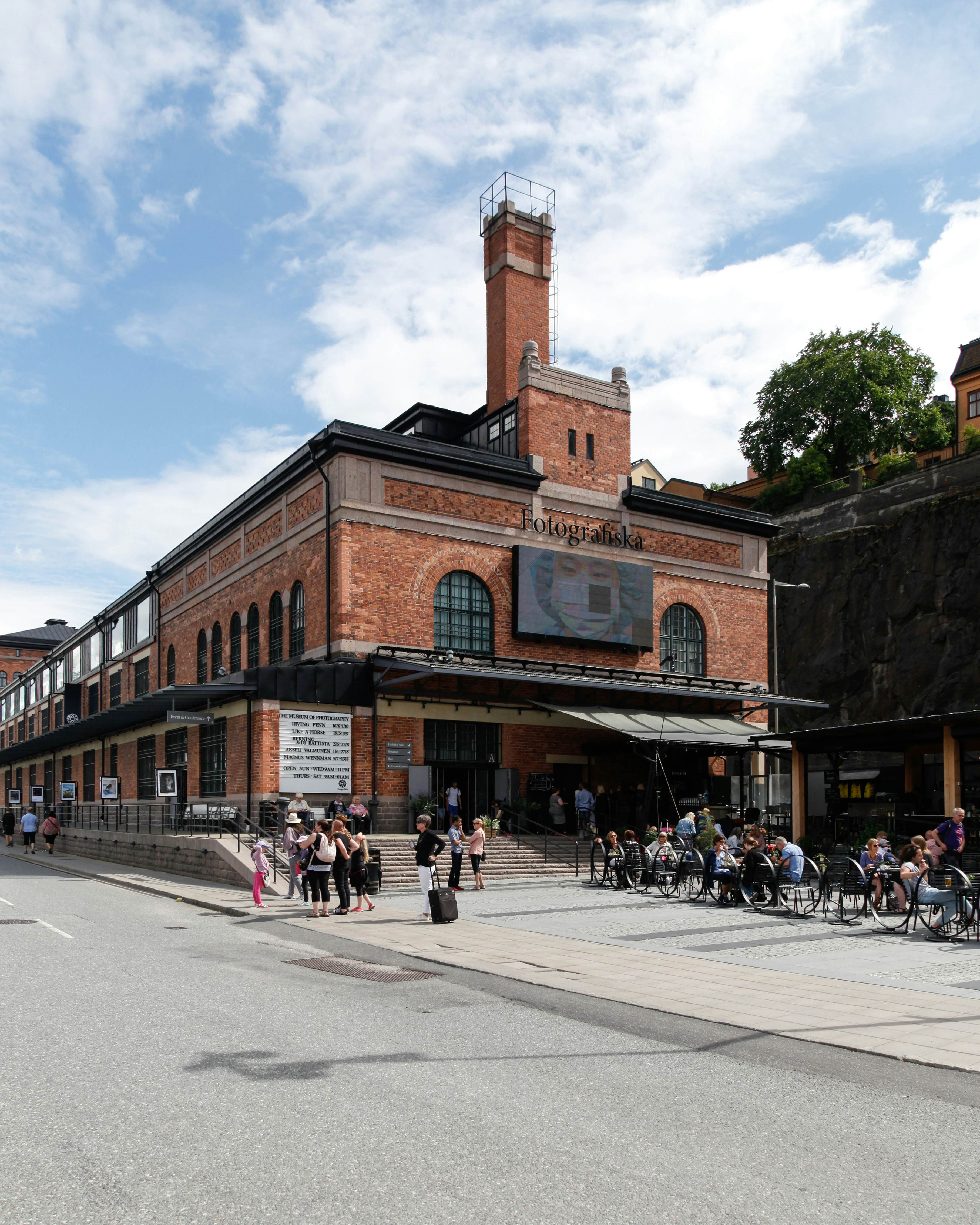 A redbrick building with people standing in front and sitting in an outdoor cafe.