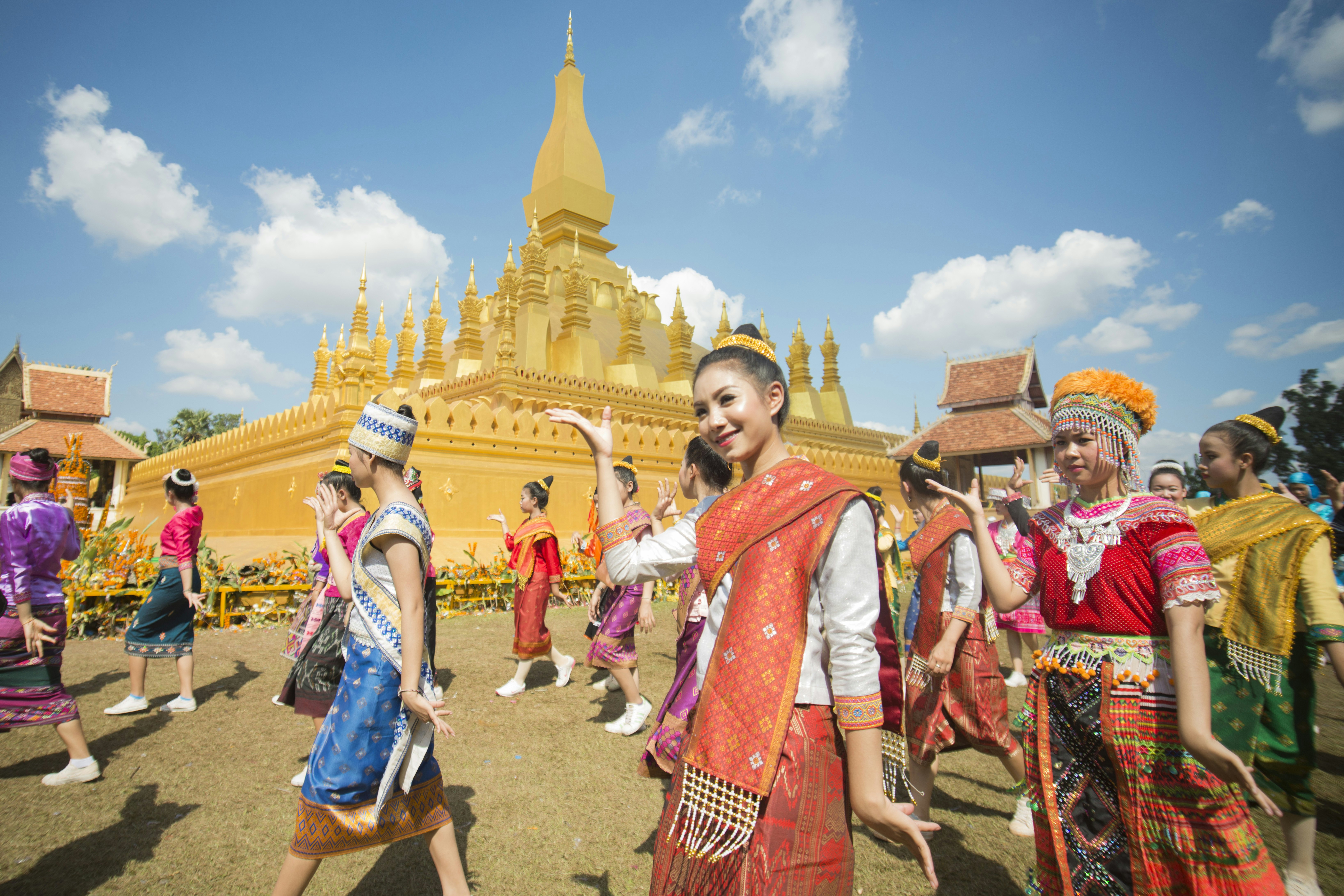 People in traditional costumes make hand gestures as they participate in a parade past a temple in a city street.