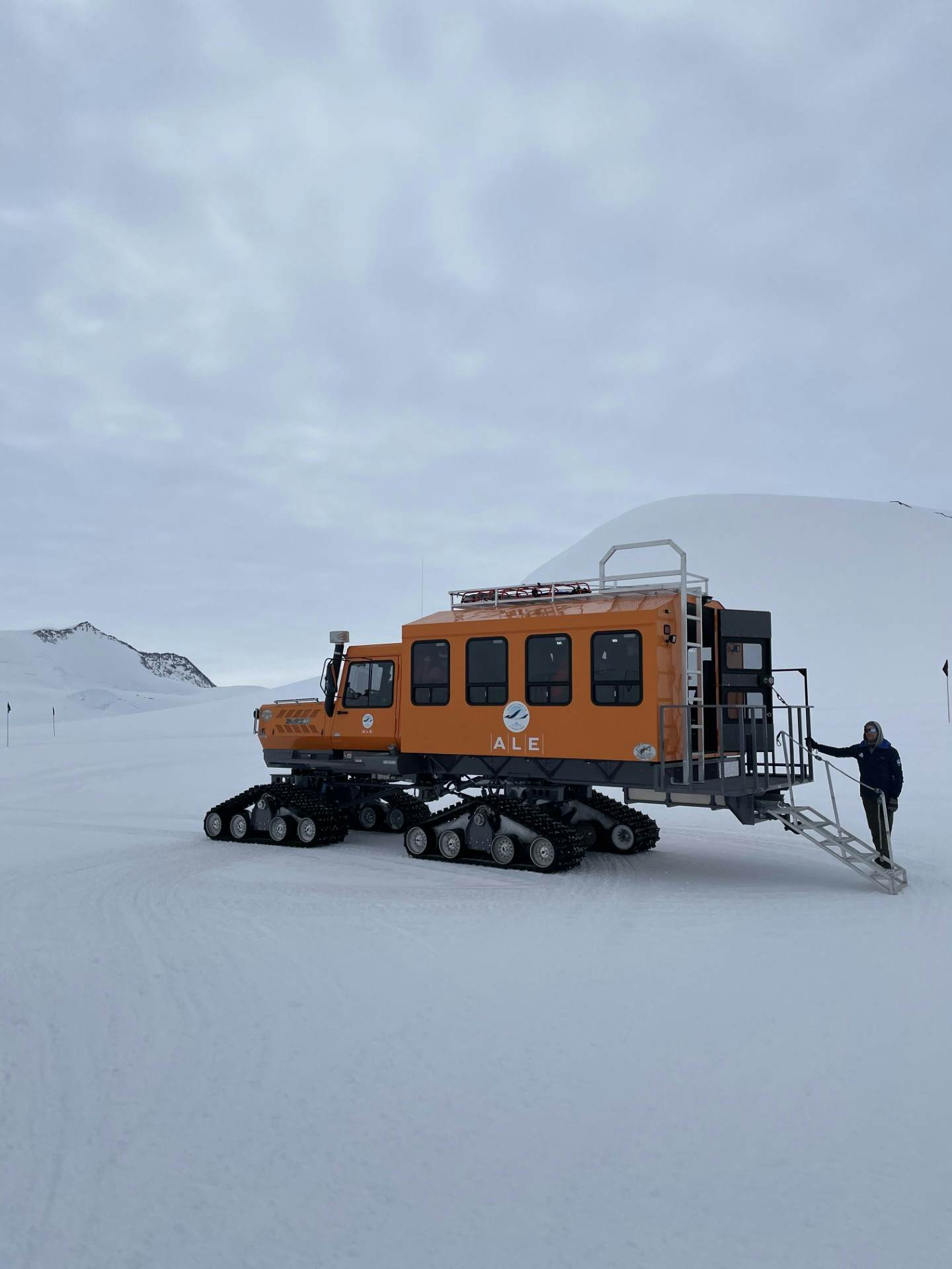 A woman stands with her arms out wide in front of a sign that reads "Union Glacier Antarctica"