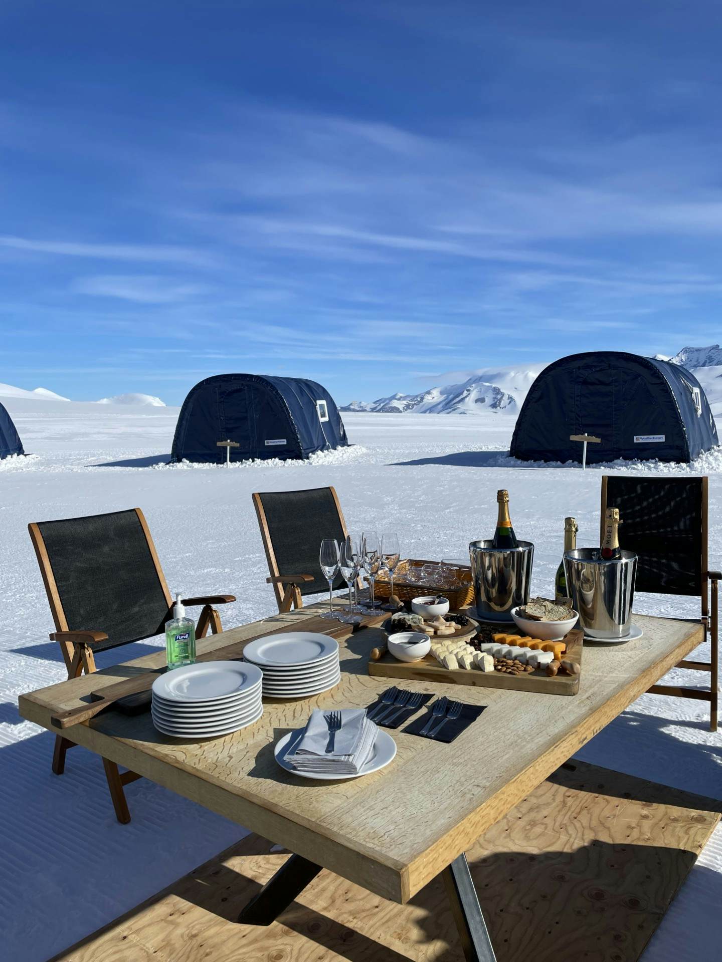 A table in the snow on a sunny day laden with Champagne bottles and sharing plates of food.
