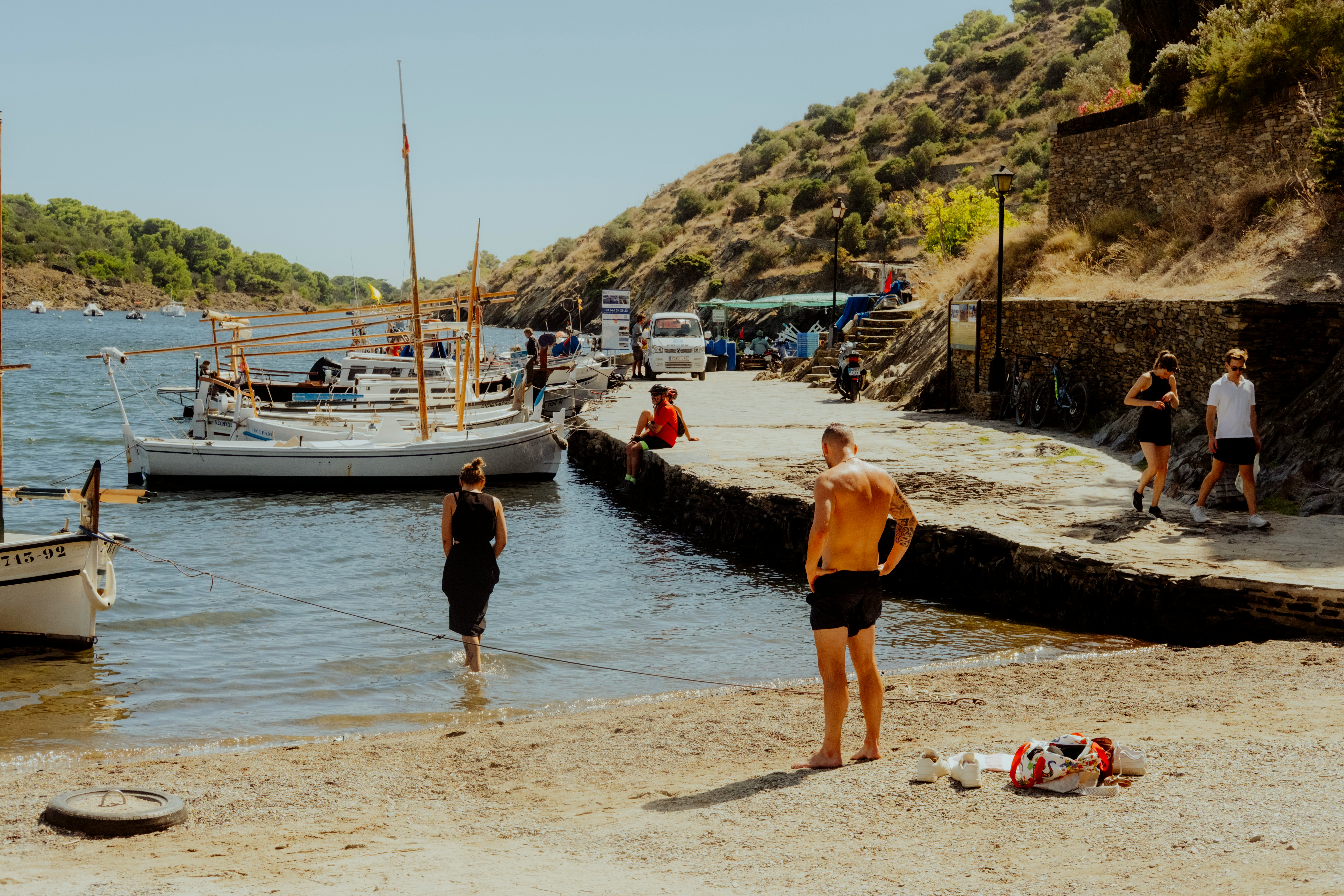 People wade into a beach near moored boats in a sheltered cove.