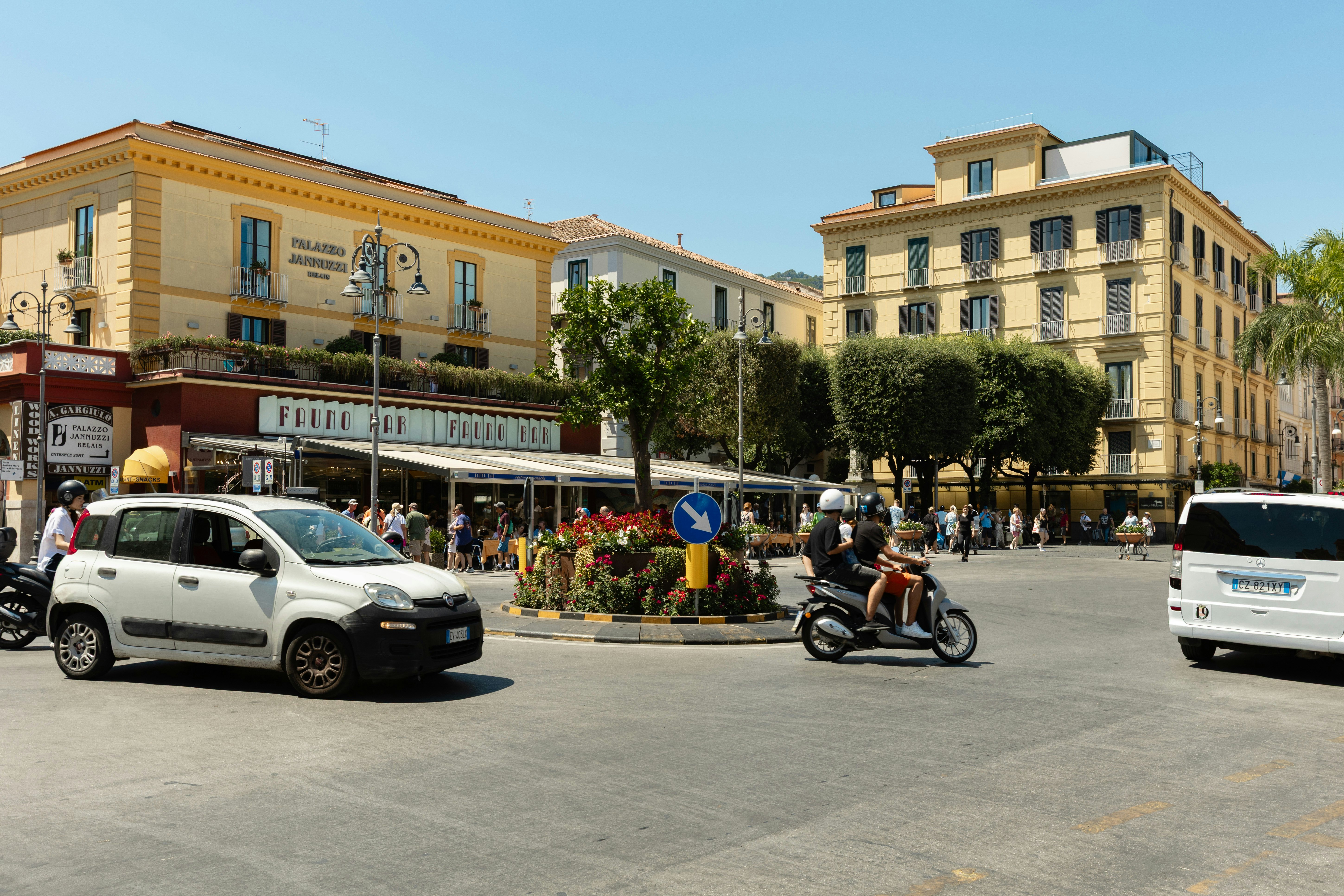 A car, Vespa and minivan make their way through a street lined with yellow and white low-profile buildings with many windows