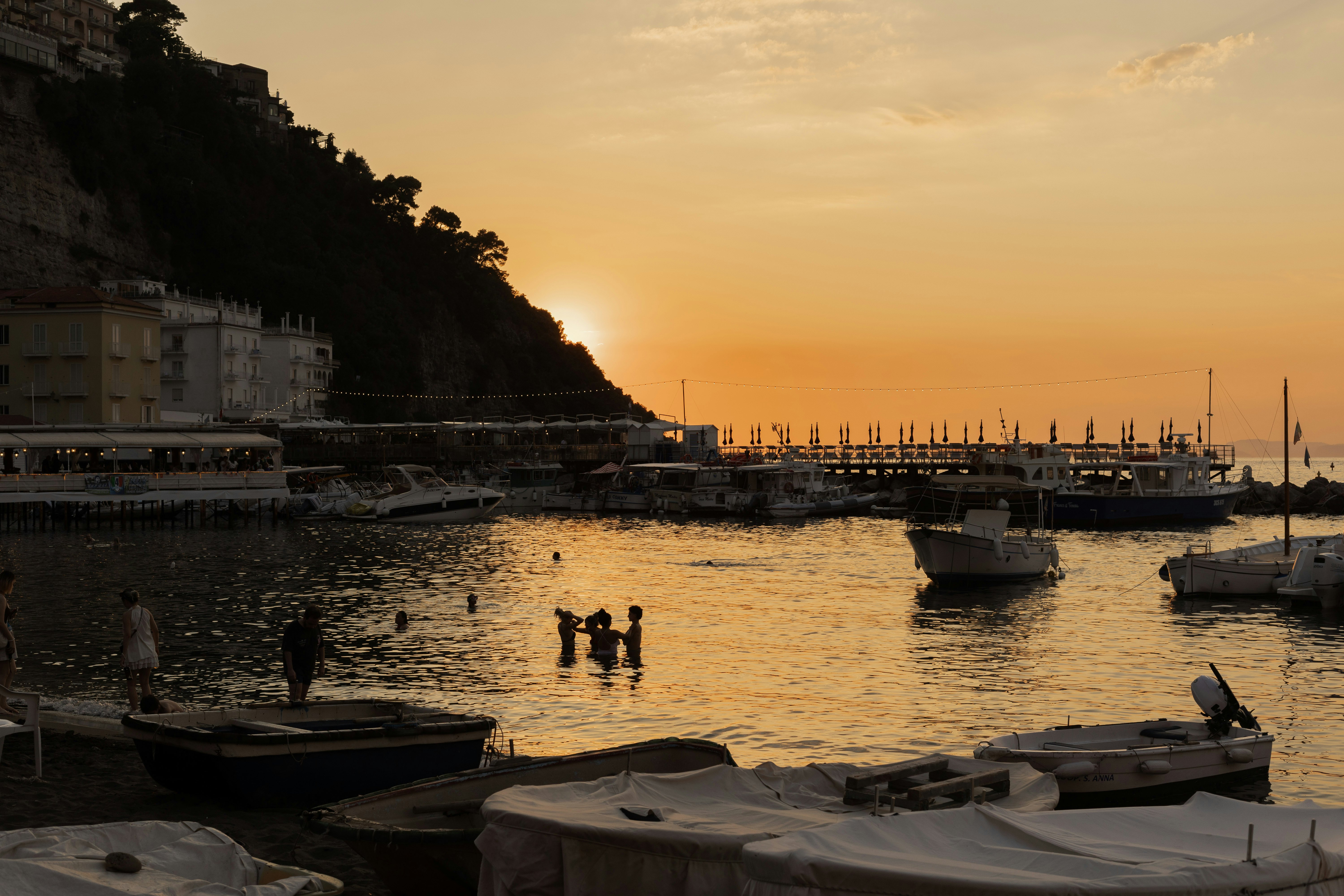 Silhouettes of people in a body of water near boats and a dock at sunset