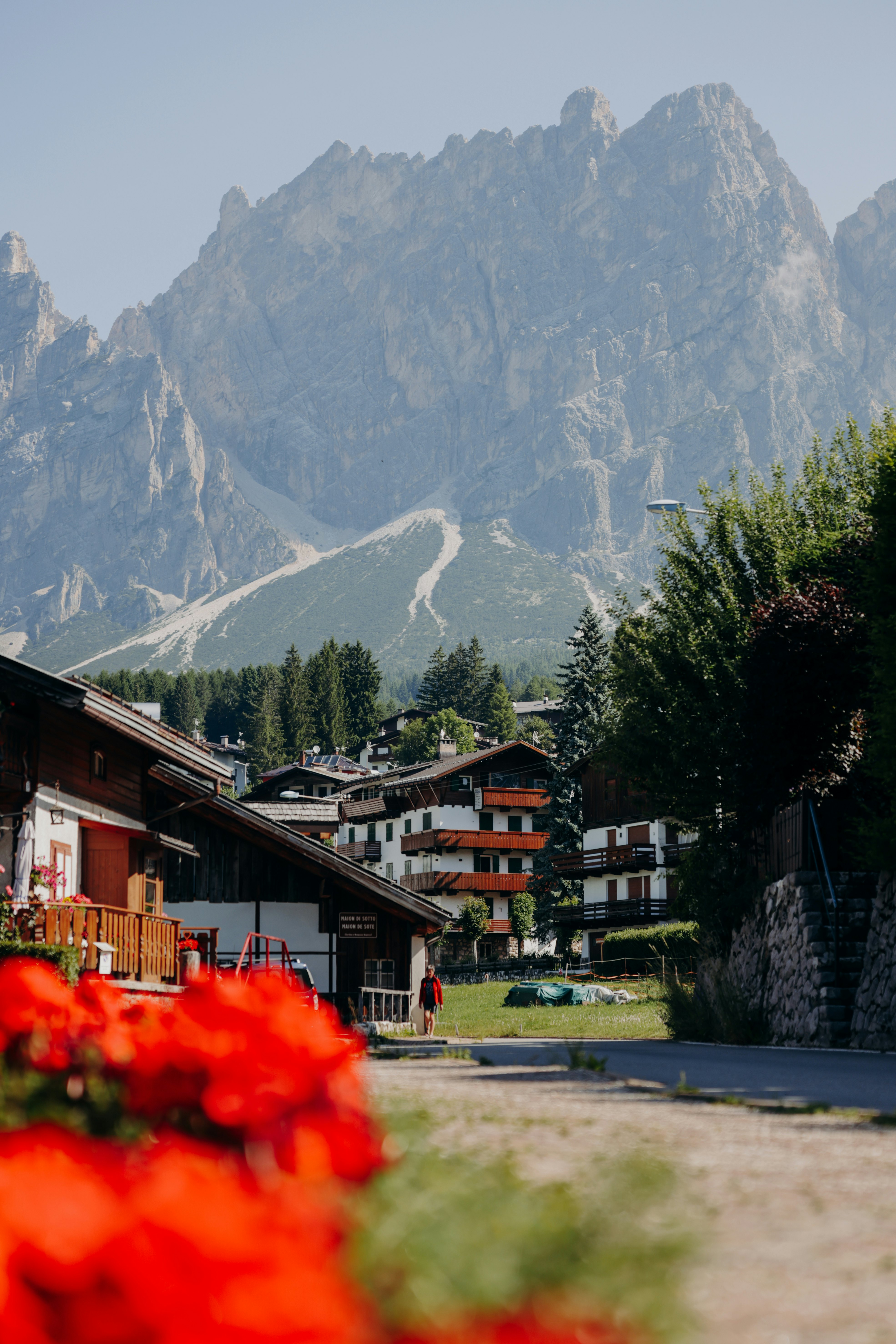 A small town in the shadow of mountains on a summer's day, with bright red flowers filling flower boxes outside chalet-style buildings.