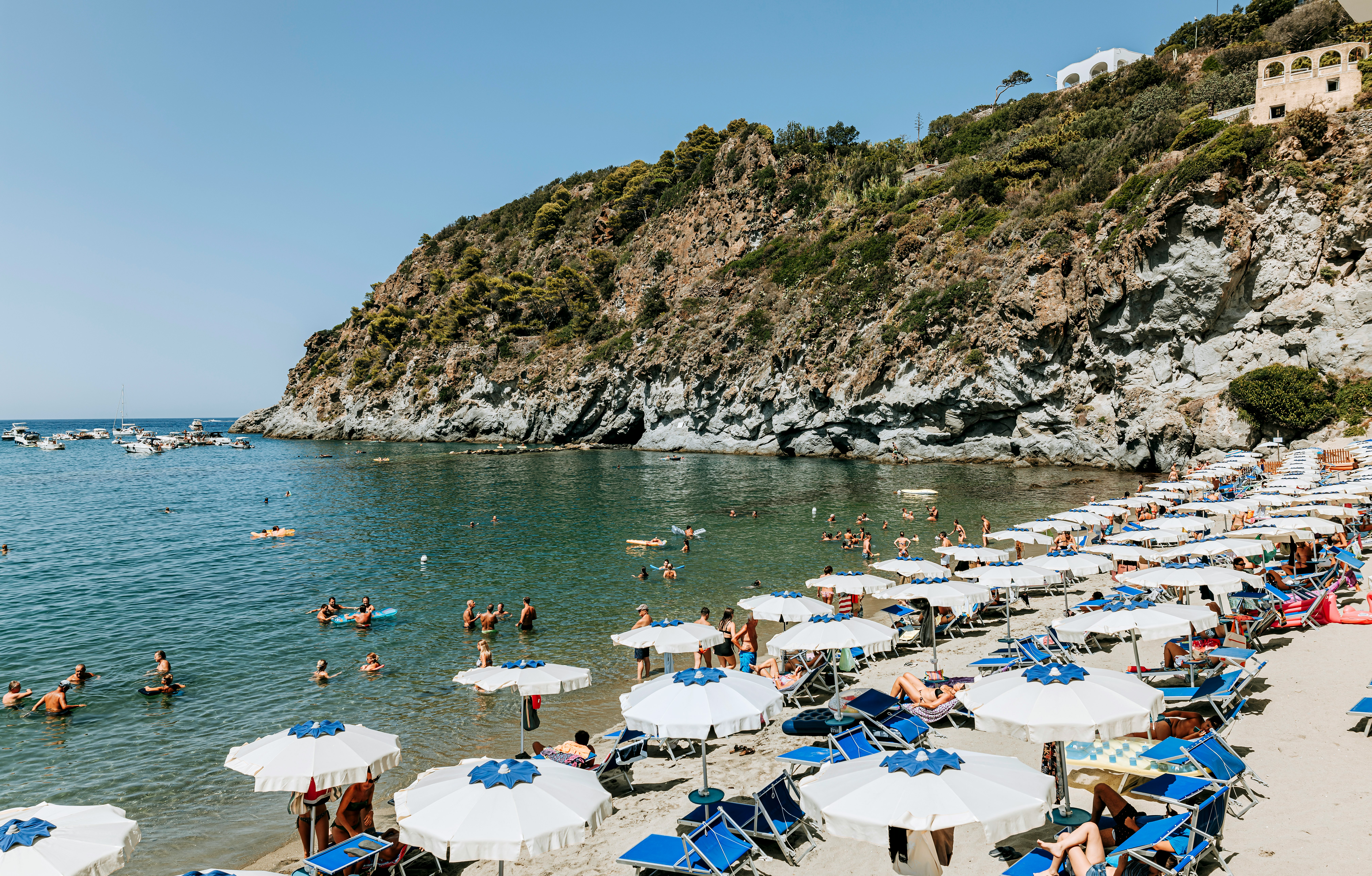 A beach is filled with sun loungers and umbrellas, with people swimming just offshore. A steep slope at the edge of the beach leads to the water.