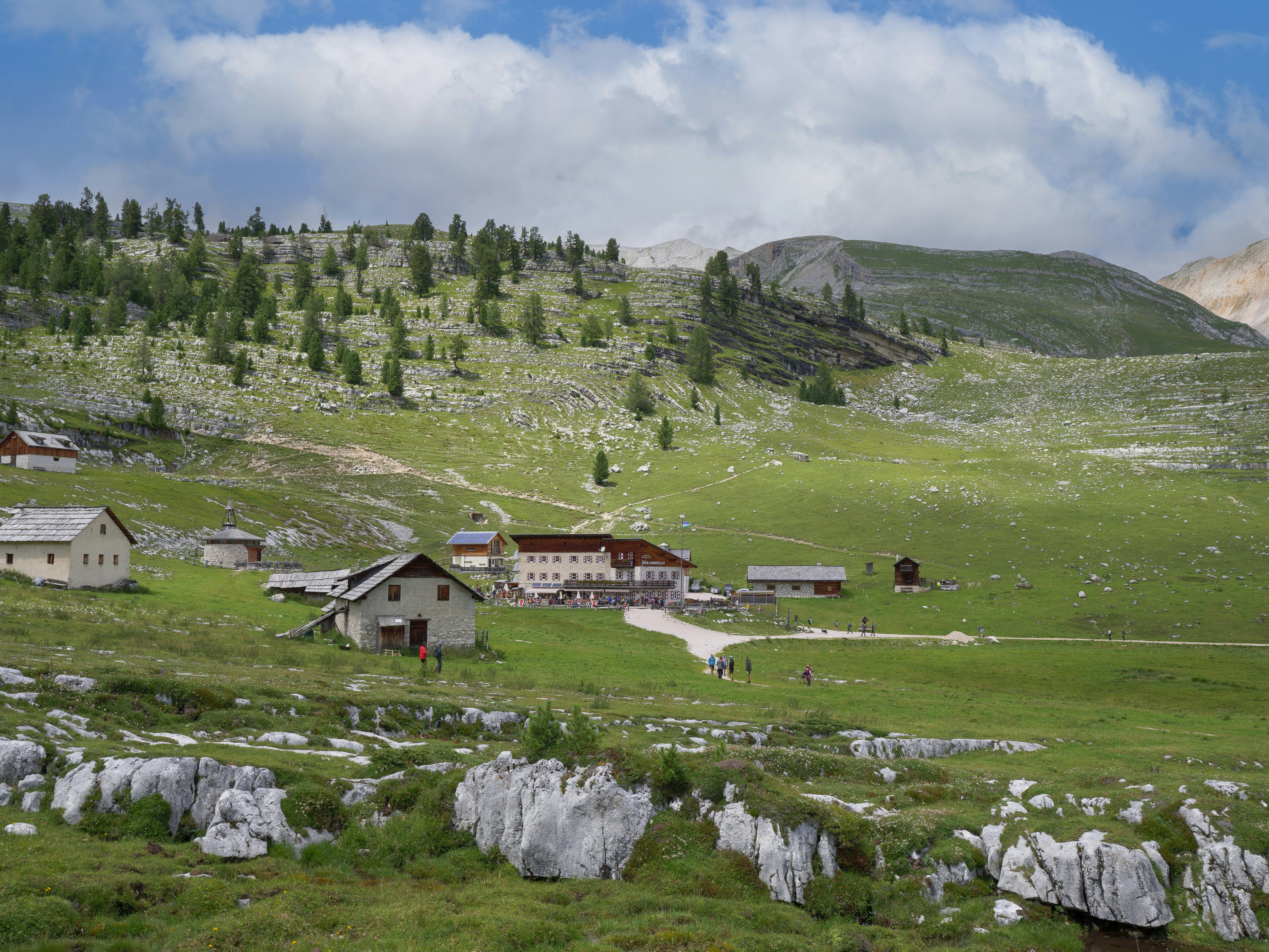 Hikers follow a trail through green fields on a mountain plateau to a series of huts and a a large chalet-style building.