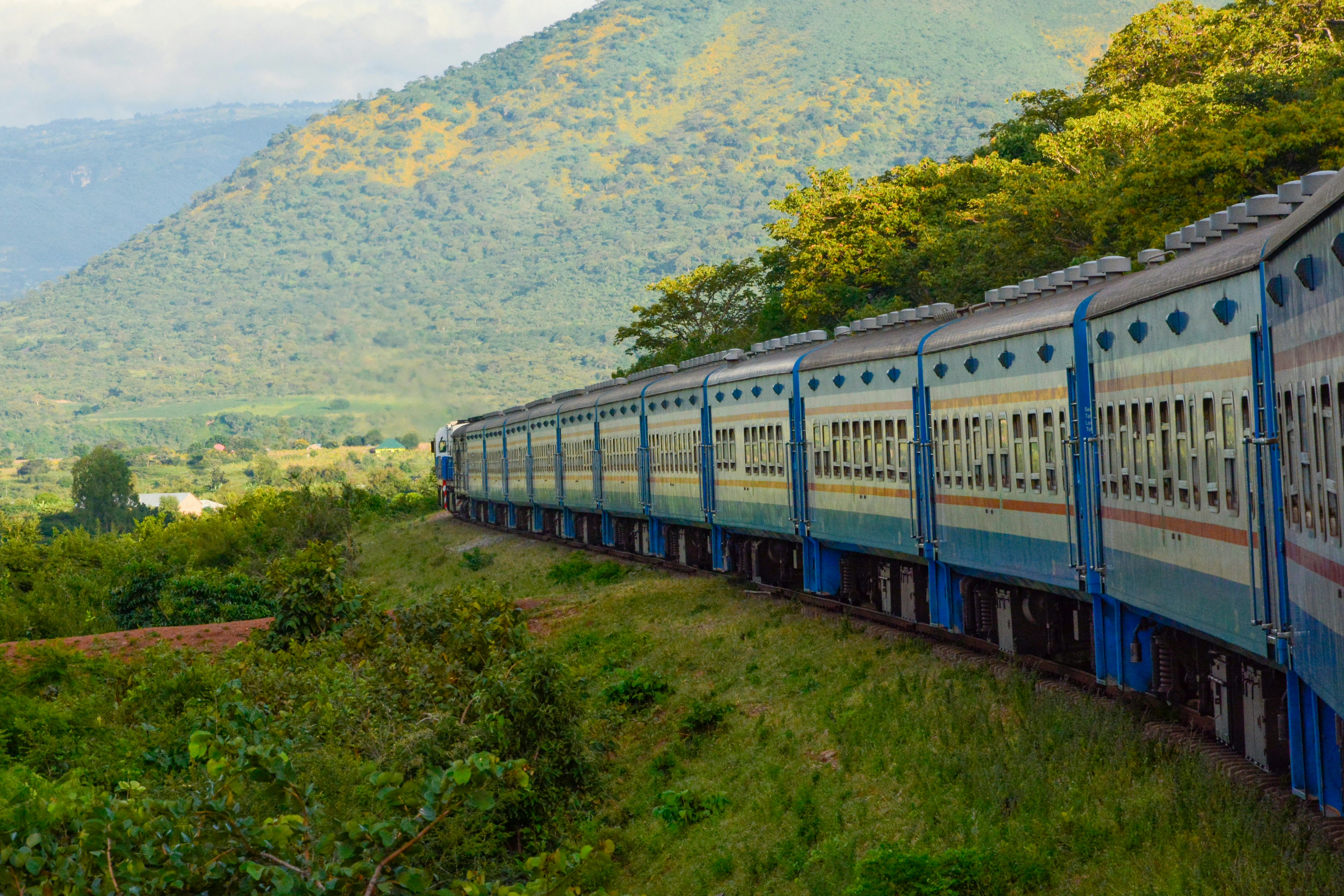 A blue and white train travels along tracks through a rural landscape of grasslands and wooded hillsides.